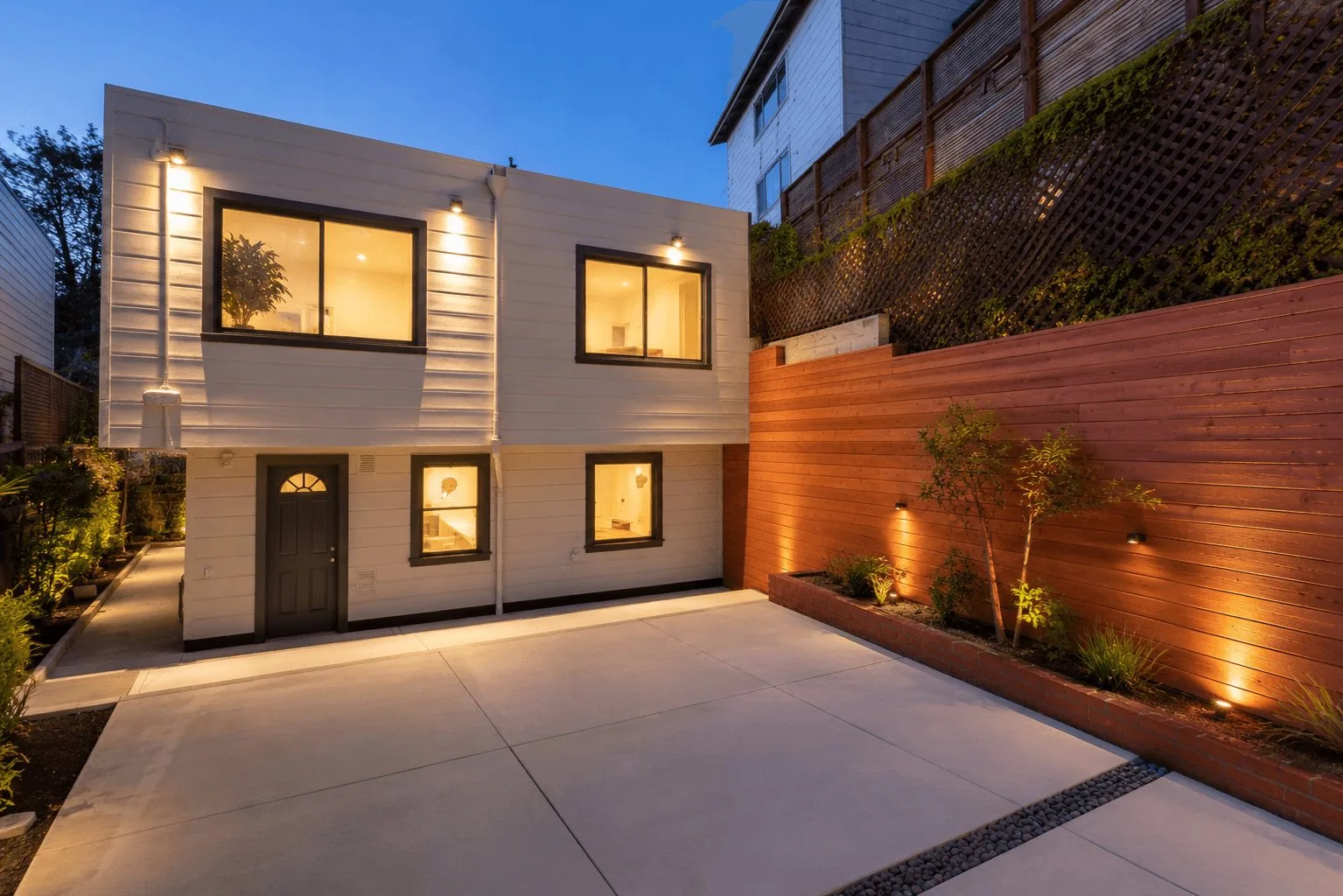 A converted garage ADU at dusk, below a two-story house, with a new patio and cedar fence