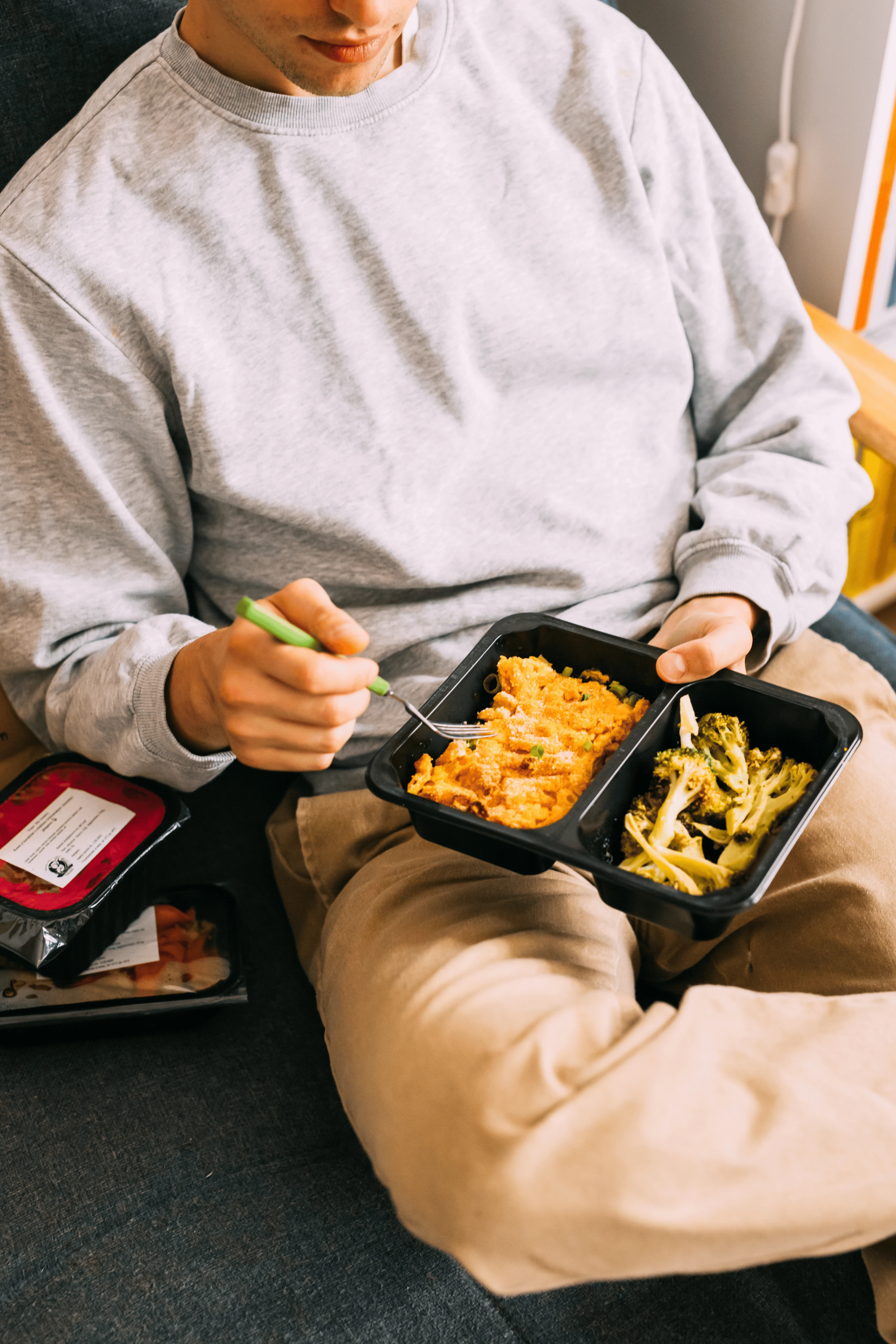 Mężczyzna na kanapie je wegański obiad z cateringu Mihiderka — w czarnym pojemniku danie z makaronu z brokułami, obok kolejne pudełko cateringowe z etykietą i opisem składu