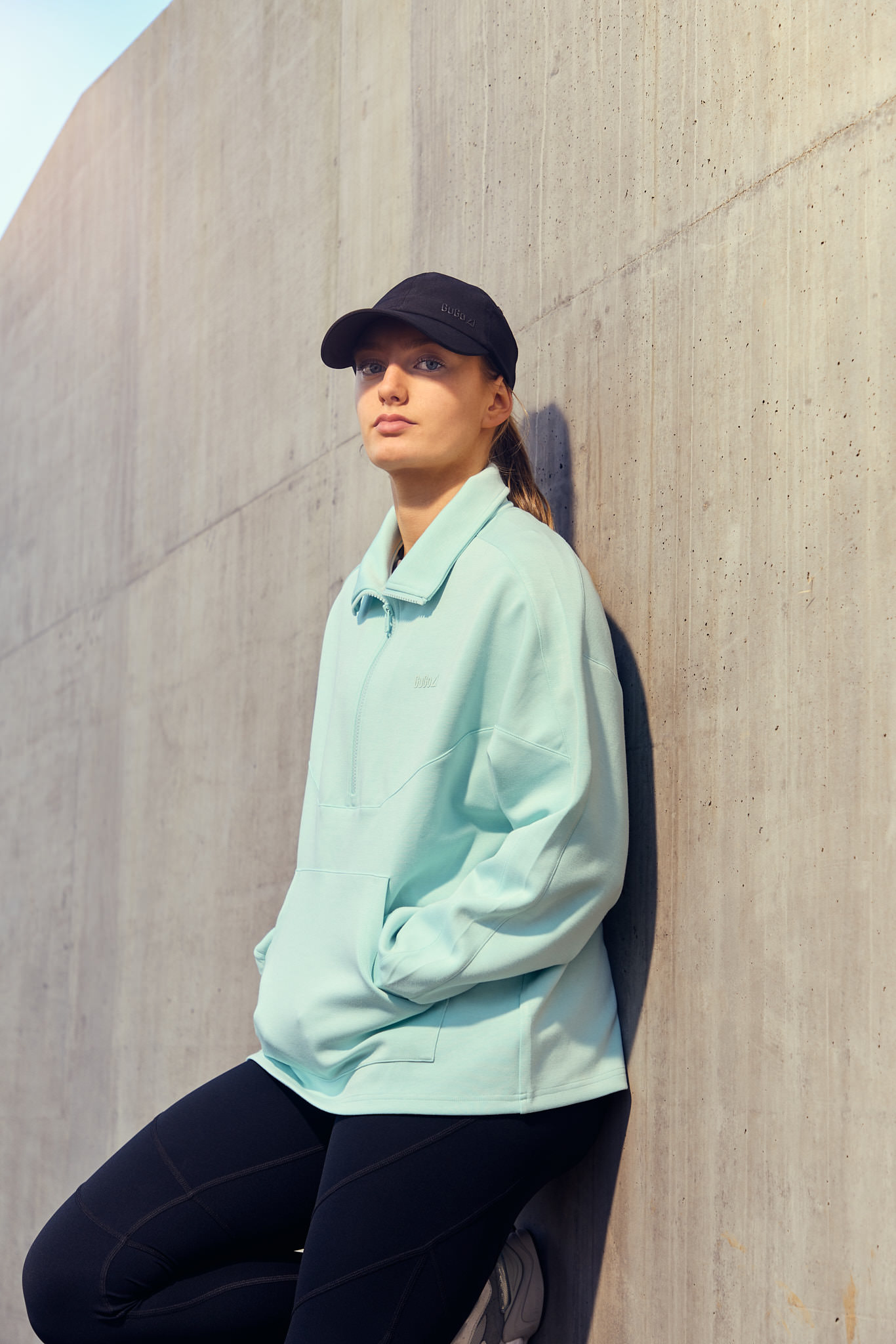 A person in a pastel green jacket and cap stands casually against a light-colored wall.