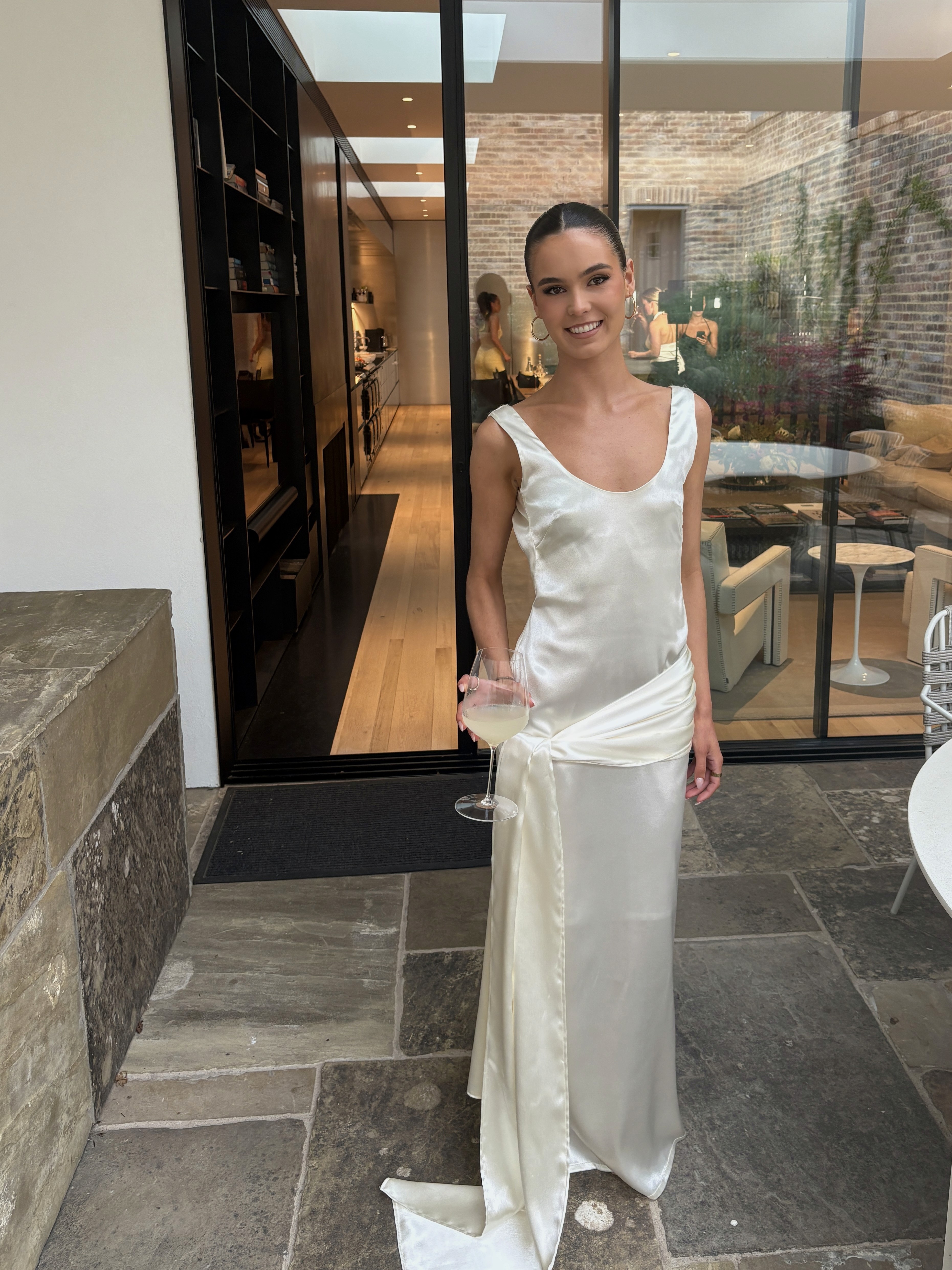 Floor-length ivory satin slip dress with scoop neckline, sleeveless cut, and asymmetrical draped sash detail at the hip