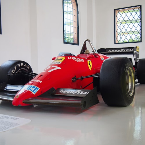 מכונית מירוץ של פורמולה 1 בצבע אדום עם צמיגי גודייר ותיחום של פרארי, מוצגת בחדר מואר היטב עם חלונות קמורים.