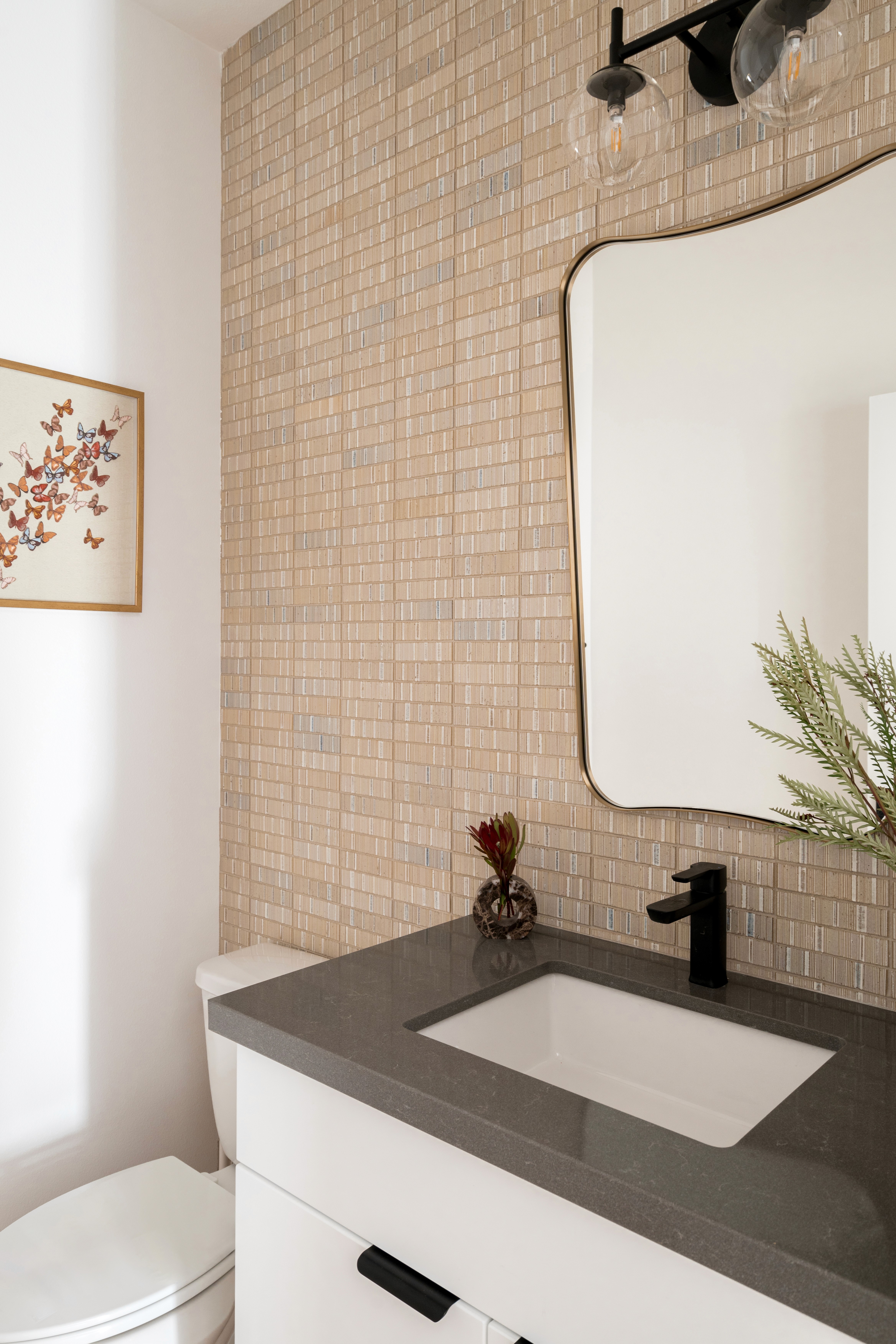Beige, textured wall paper with rectangular pattern, rounded vanity mirror, dark grey countertop, and black faucet