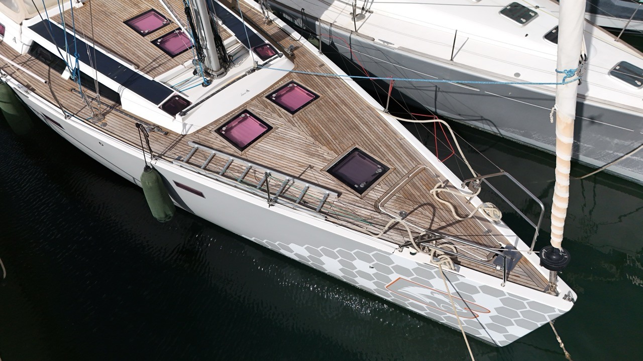 Close aerial drone view of the Hanse 470e foredeck and bow, highlighting the full EcoDeck teak-tone composite deck installation with multiple flush purple-tinted deck hatches, bow anchor platform, and distinctive grey hexagonal hull graphic wrap