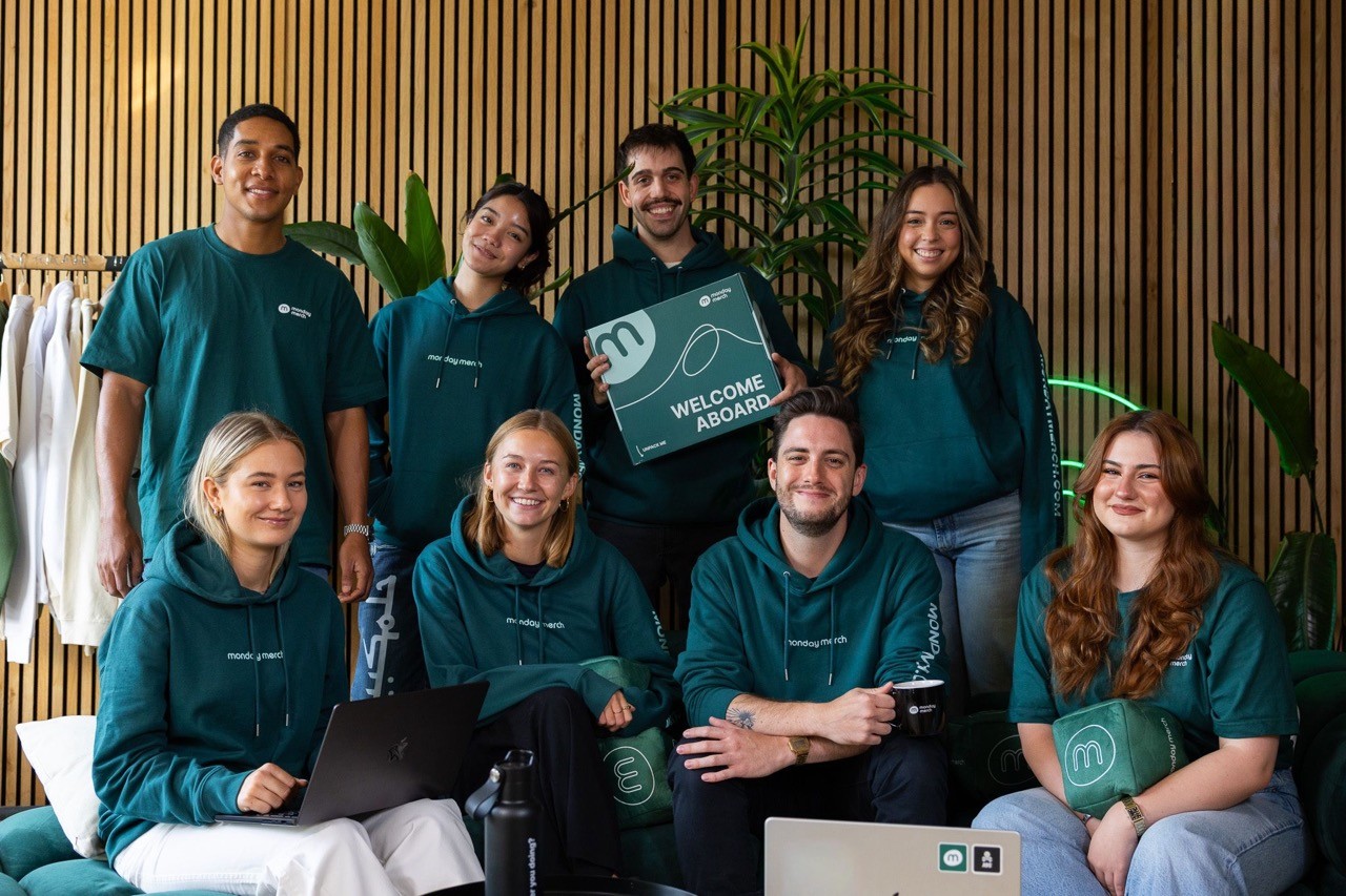 Five people wearing matching green tops pose together in a cozy indoor setting with plants in the background.
