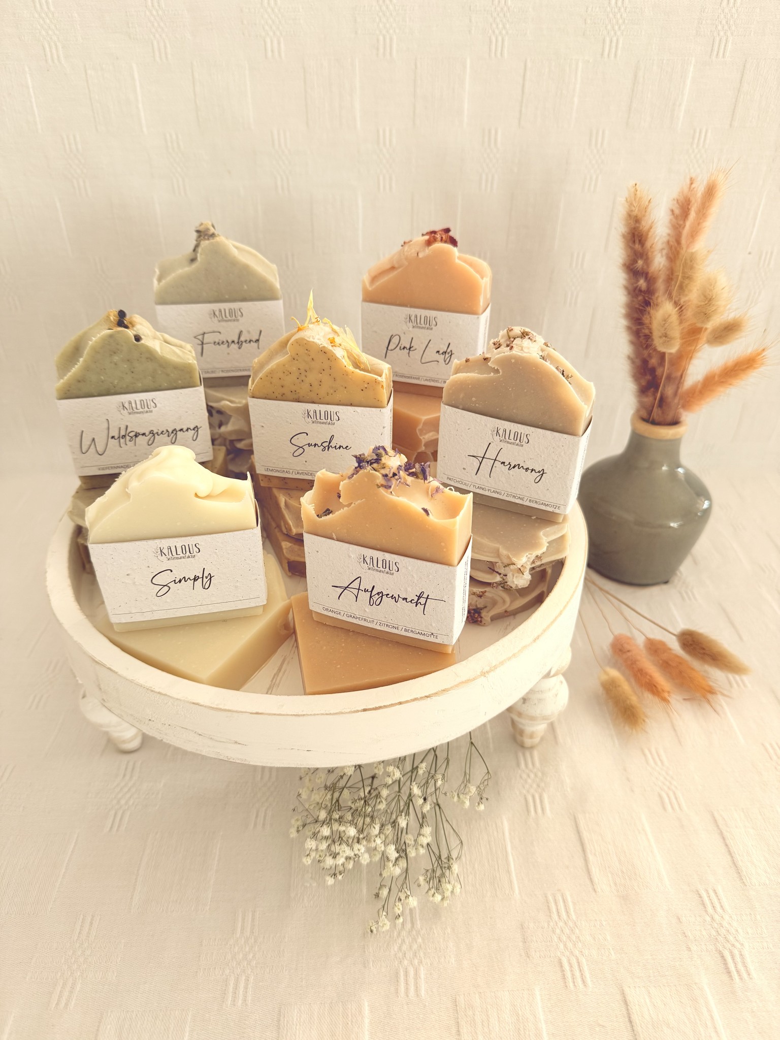 Handgemachte Naturseifen von Kalous auf einem runden Holzpodest. Sorten wie "Waldspaziergang“ oder "Harmony", liebevoll arrangiert mit Trockenblumen im Hintergrund.