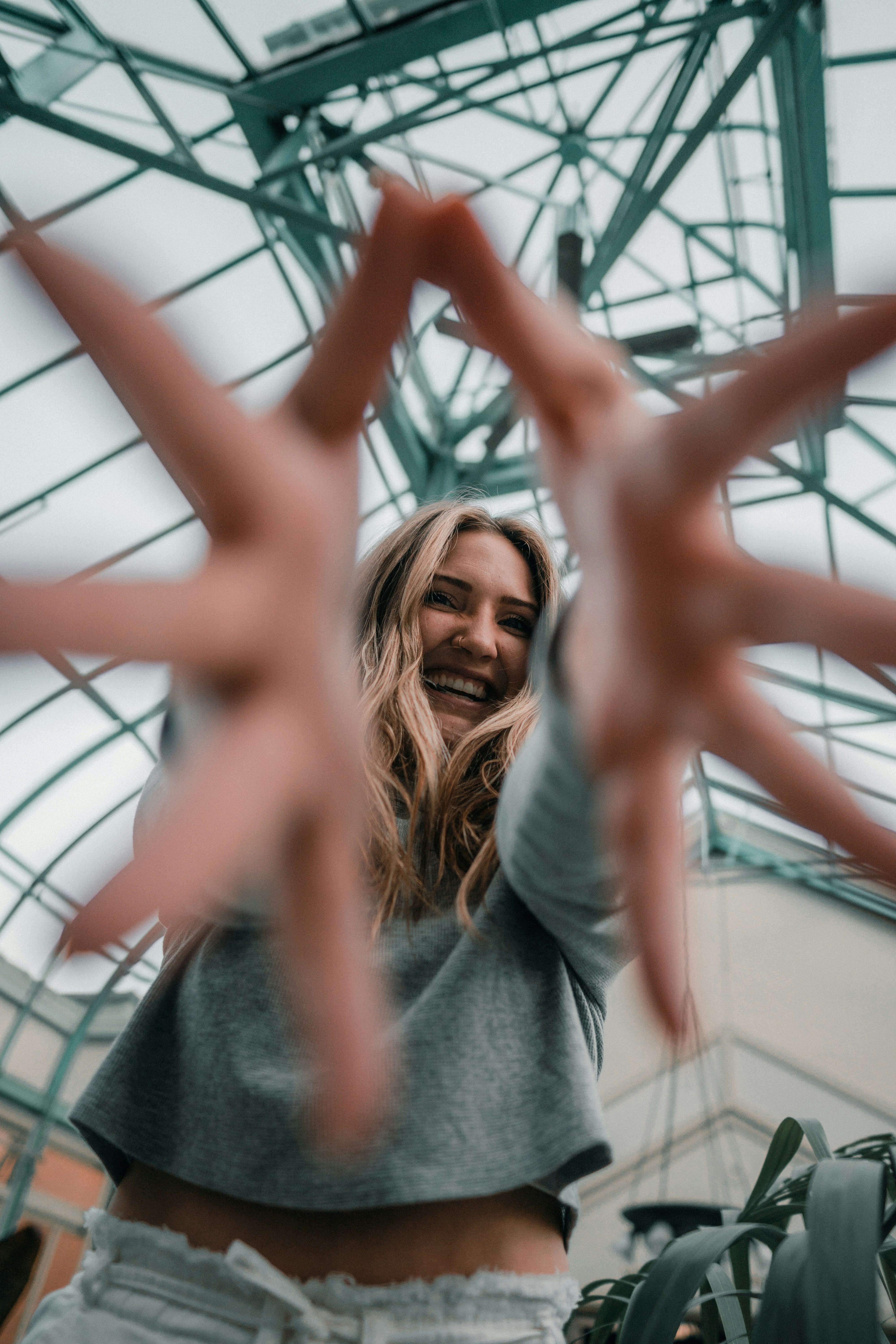 A woman reaching toward the camera inside a glass greenhouse with metal framing
