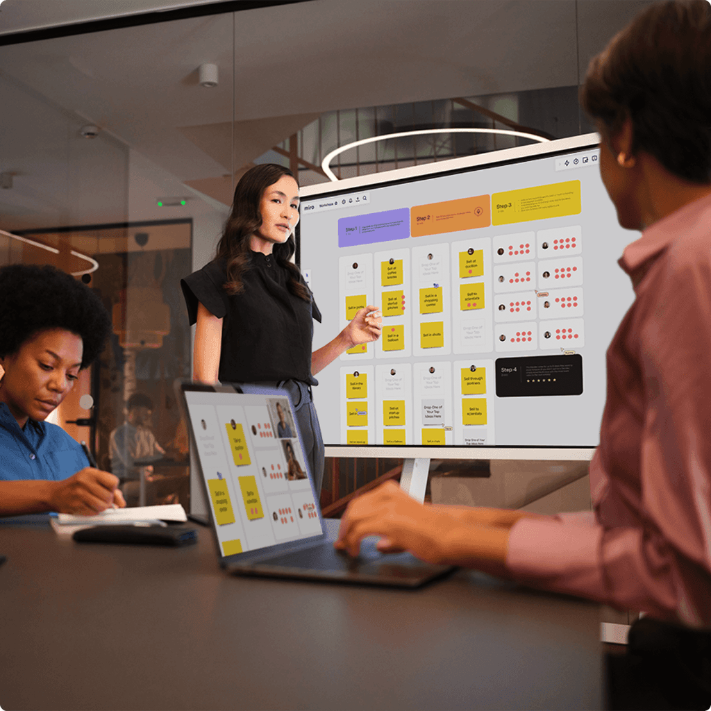 In a modern conference room, three professionals engage in a collaborative discussion while using a digital whiteboard displaying a Miro workspace filled with colorful sticky notes and diagrams.