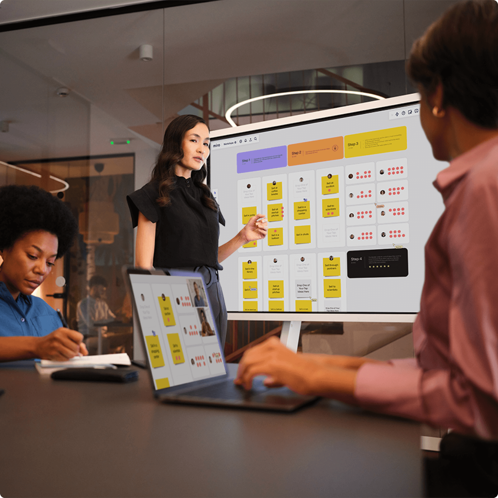In a modern conference room, three professionals engage in a collaborative discussion while using a digital whiteboard displaying a Miro workspace filled with colorful sticky notes and diagrams.