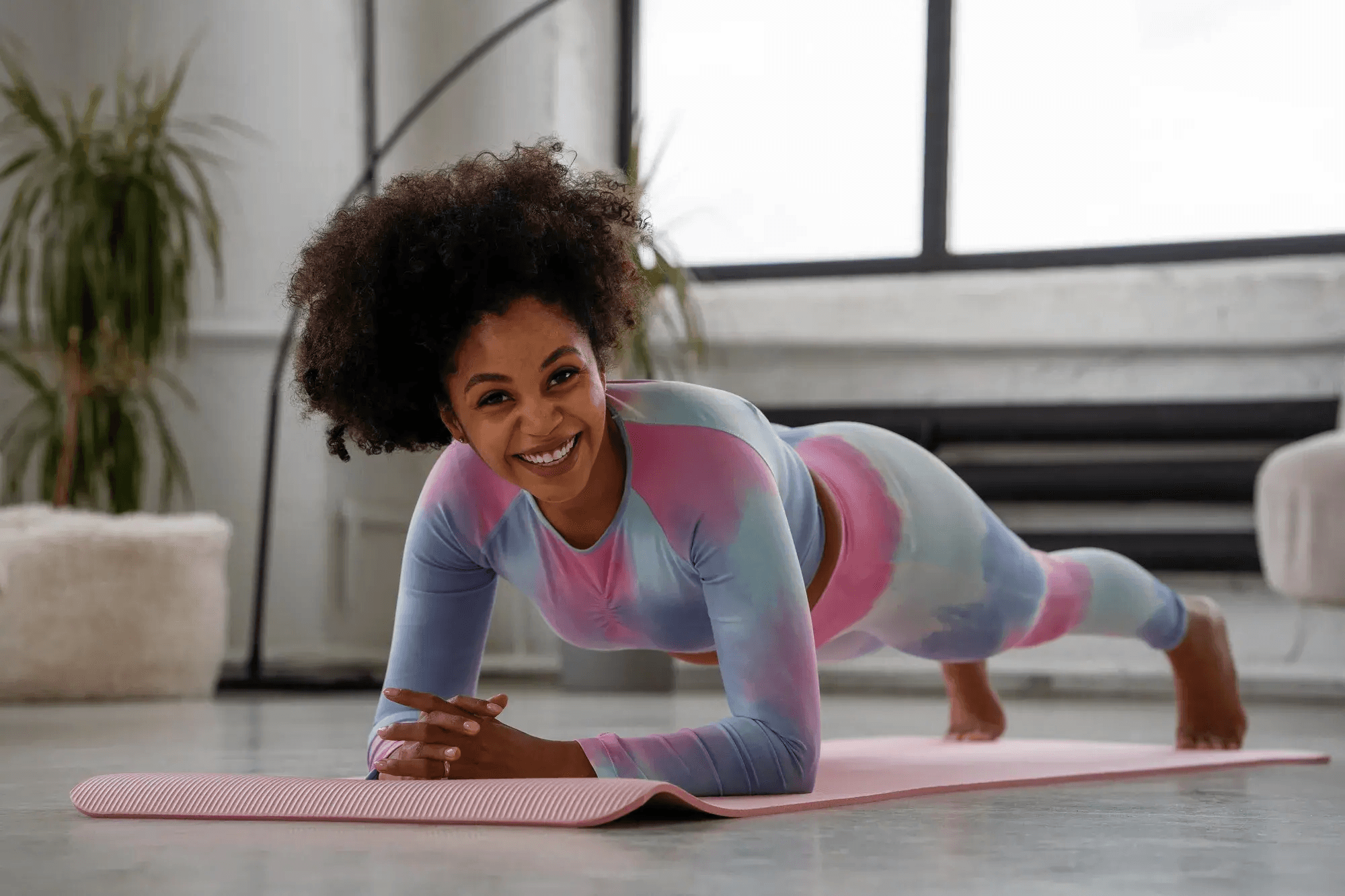 A person in colorful tie-dye athletic wear exercises on a pink yoga mat, performing a plank with a confident smile in a brightly lit living room.