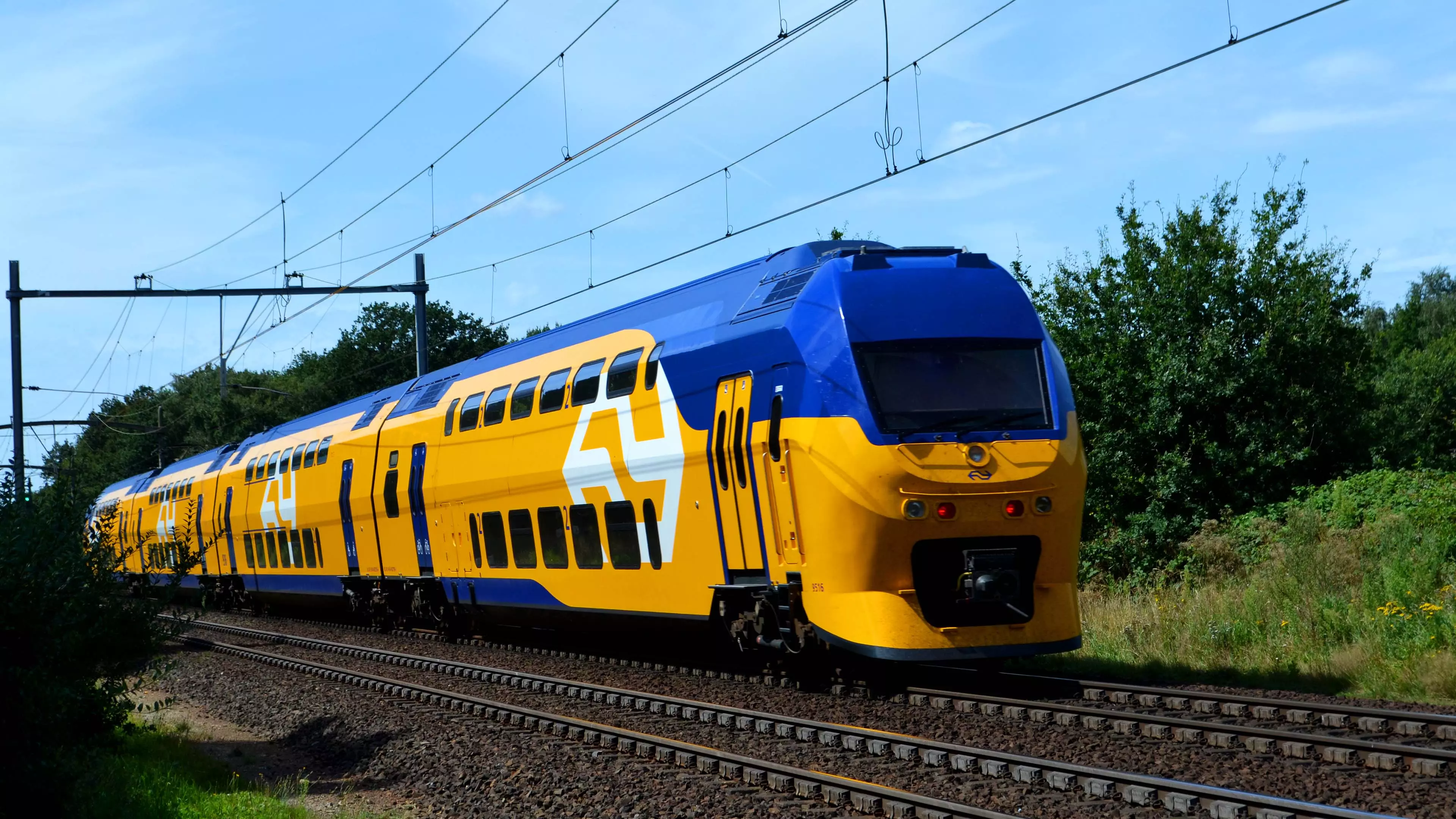 Moderne NS-trein in opvallende gele en blauwe kleuren rijdt over het spoor, met een helderblauwe lucht op de achtergrond. De afbeelding toont de innovatieve en herkenbare stijl van de Nederlandse Spoorwegen, wat de efficiëntie van het treinvervoer in Nederland benadrukt. Deze trein is een voorbeeld van duurzaam openbaar vervoer, ideaal voor zowel dagelijkse pendelaars als toeristen die de schoonheid van Nederland willen verkennen.