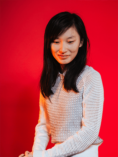 A woman with long, dark hair stands against a solid red background. She wears a light-colored, textured sweater over a white collared shirt. Her arms are crossed in front of her, and her gaze is directed slightly downward and to her right. She has a gentle smile on her face.