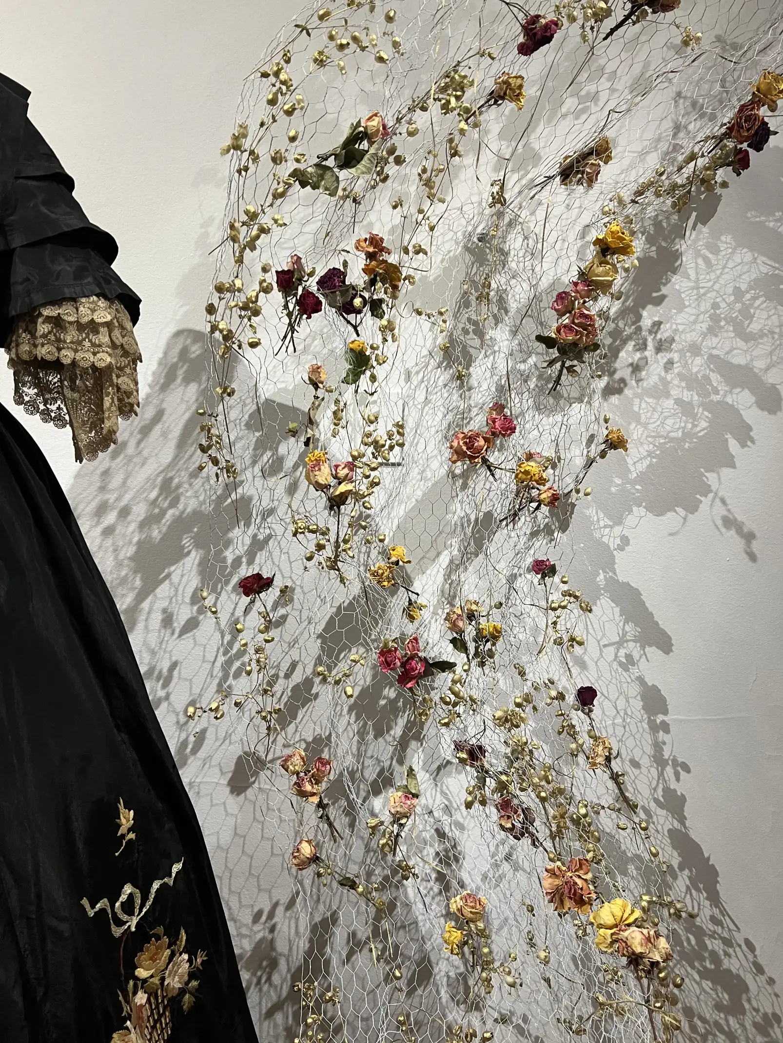 Instalación de flores secas y malla metálica junto a vestido bordado para muestra artística que une historia y naturaleza.