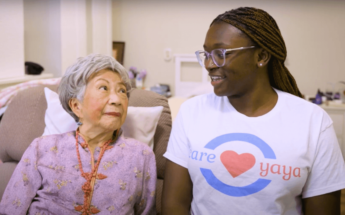 Older asian woman and young black woman in CareYaya t-shirt smile at each other while sitting on a sofa together