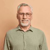 A smiling older man with glasses and a beard, wearing a green shirt against a light beige background.