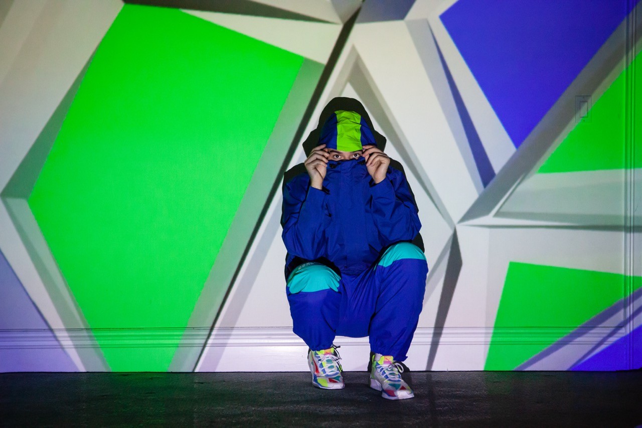 Eric Whiteback in a blue Puma tracksuit sitting in front of a geometric wall with green and blue light projections.