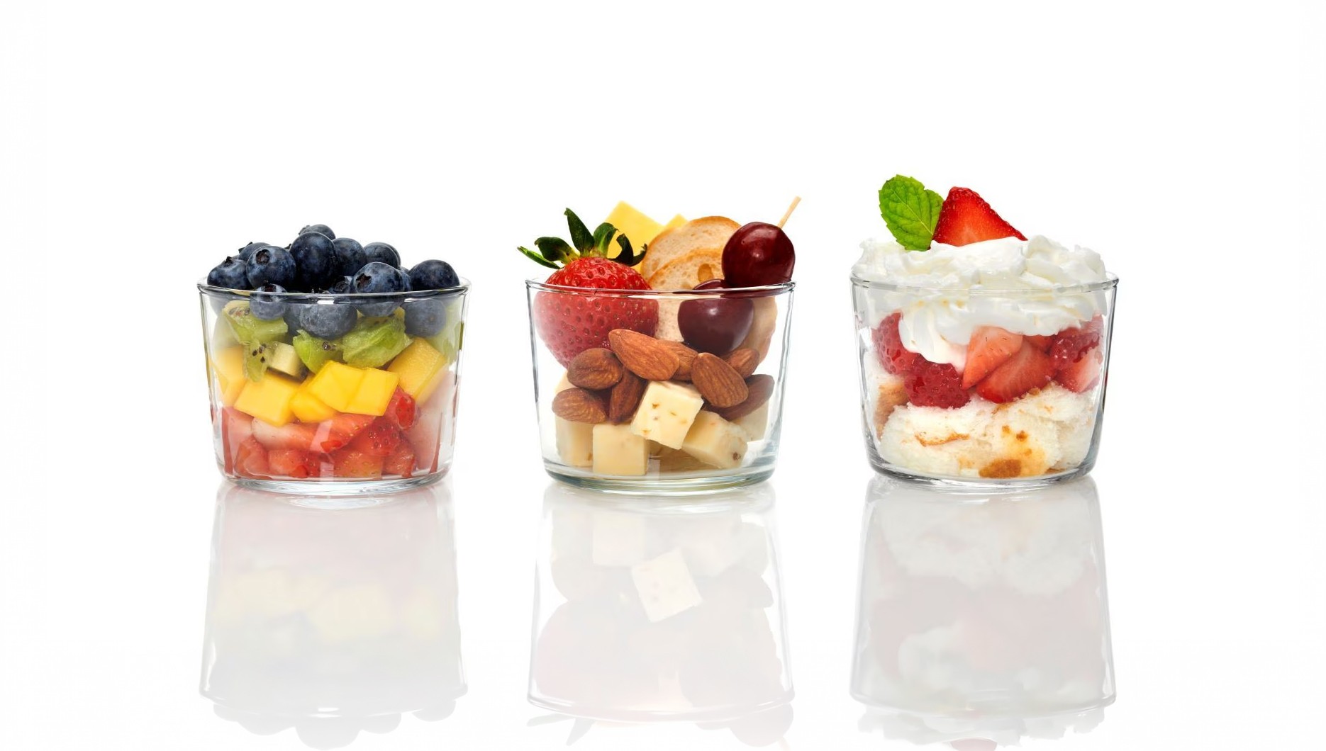 Trois verrines en verre transparent garnies de salade de fruits, fruits secs et trifle fraise chantilly, sur fond blanc