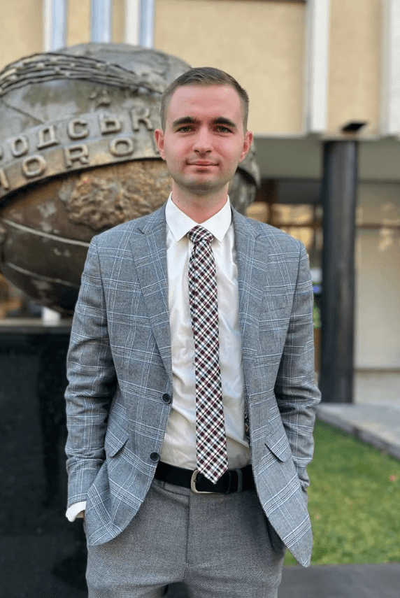 A man in a light gray suit stands confidently outside, with a decorative sphere structure in the background.