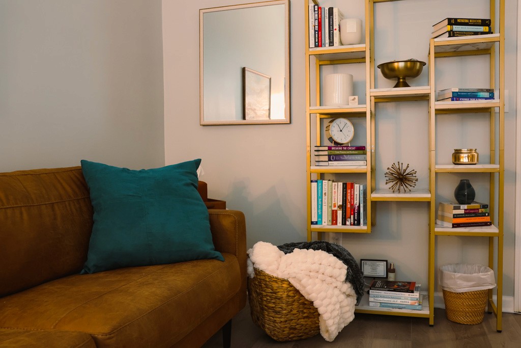 Counseling room corner with sofa, wall mirror, bookshelf, and folded blanket in a basket.