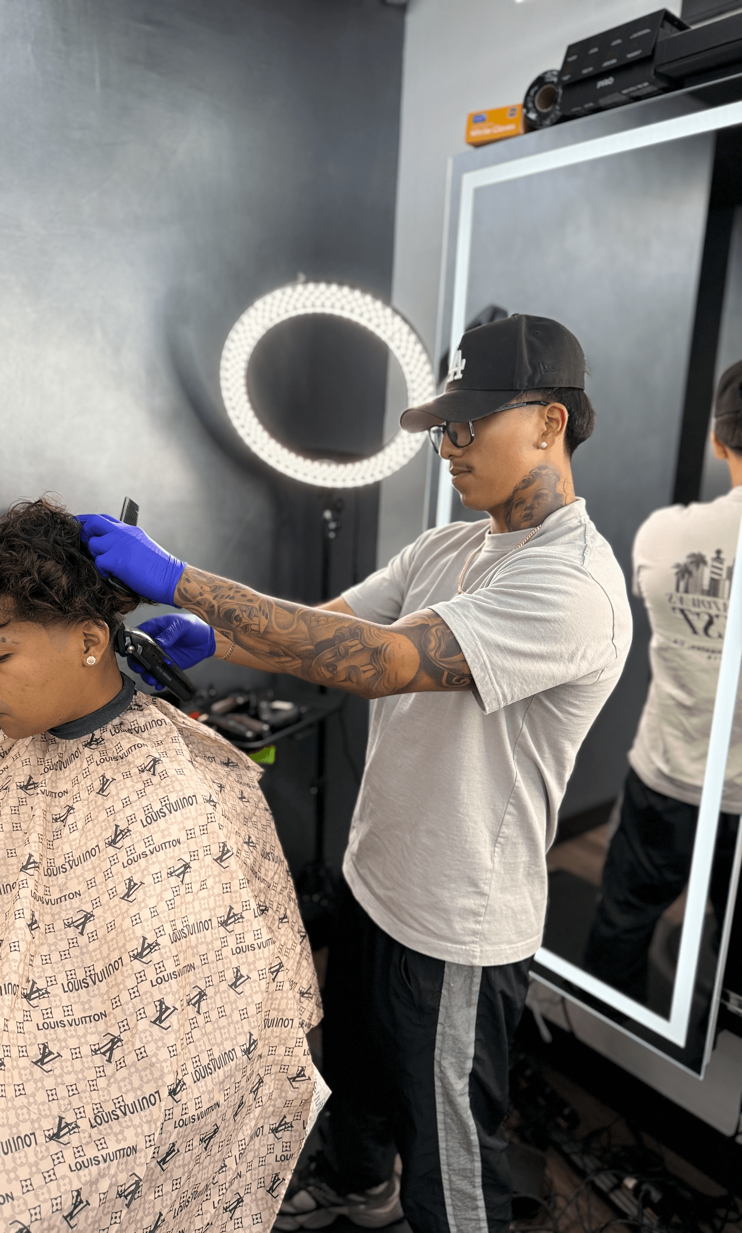 A barber shapes a client’s haircut using clippers near a bright ring light in barbershop. 