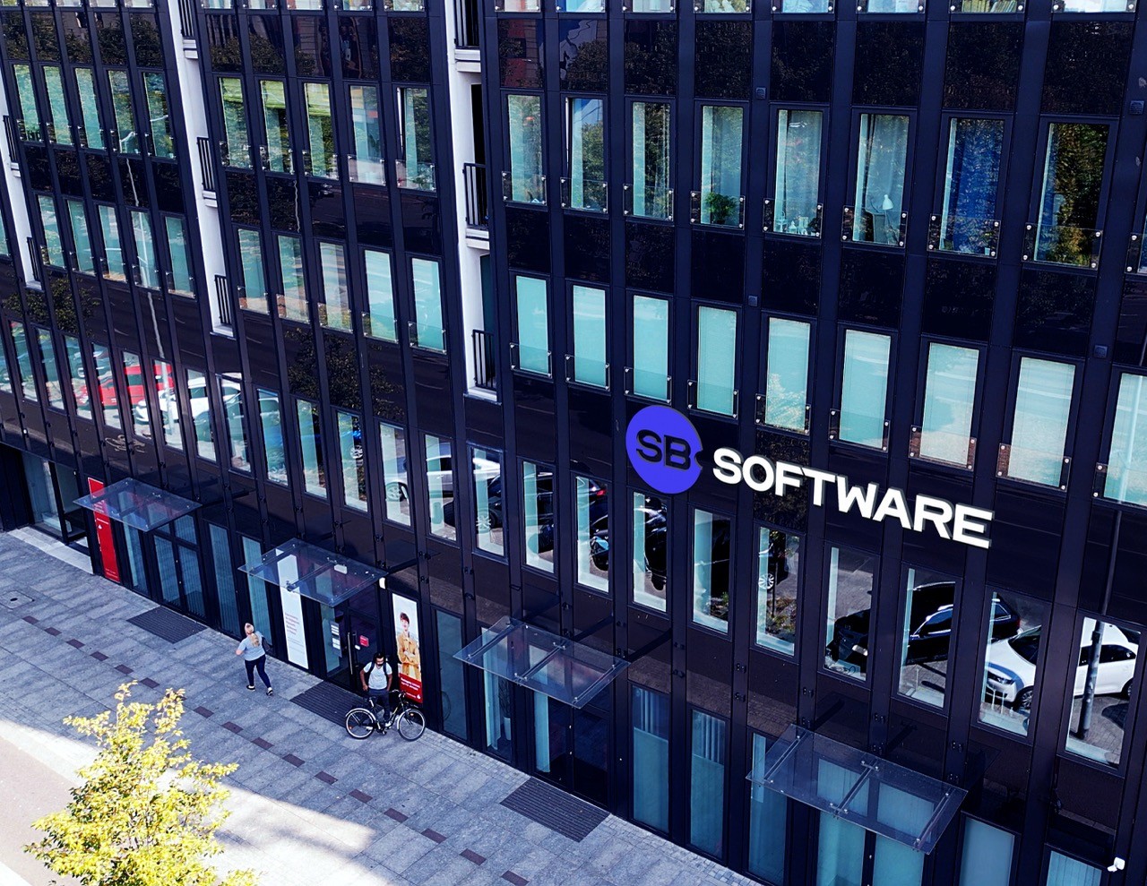 The exterior of a modern office building with large glass windows, displaying the "SB SOFTWARE" logo in white letters next to a blue circle with "SB" inside it. Reflections of cars and pedestrians are visible in the windows, suggesting an urban setting.