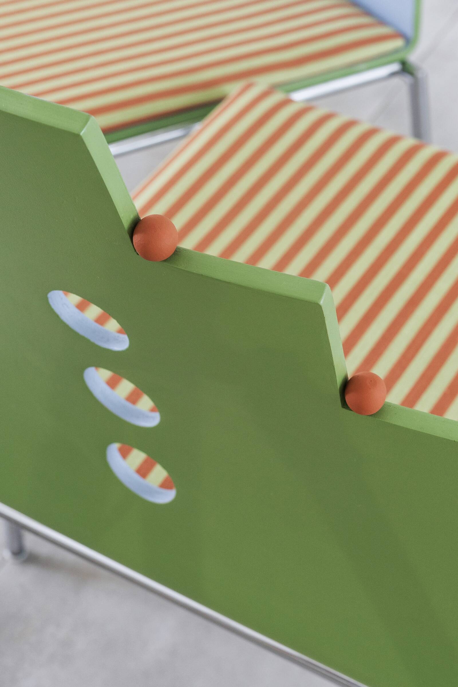 Close-up of a green wooden chair with three circular cutouts on the backrest and small orange balls at the top of the backrest, with high contrast. In the background, there are striped orange and cream patterned cushions.