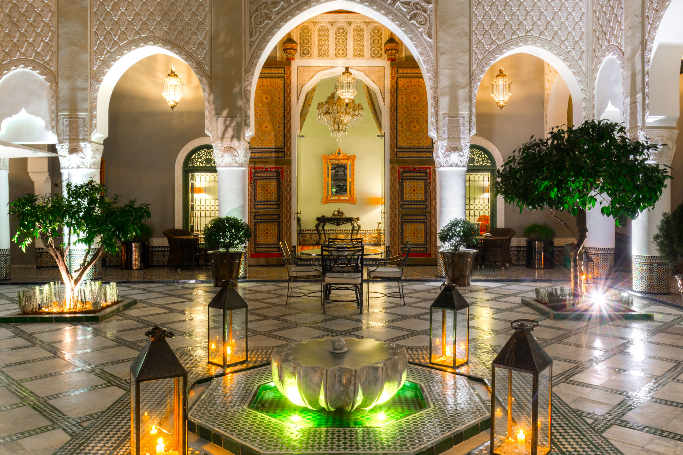 A symmetrical interior view of a palace courtyard with arched doorways, marble floors, and lit lanterns at dusk.