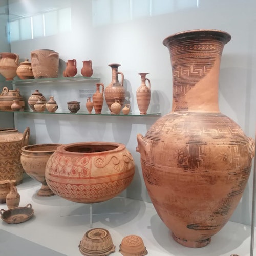 Display of ancient pottery in a museum, featuring large, ornately decorated vases and jars on shelves and a white counter.