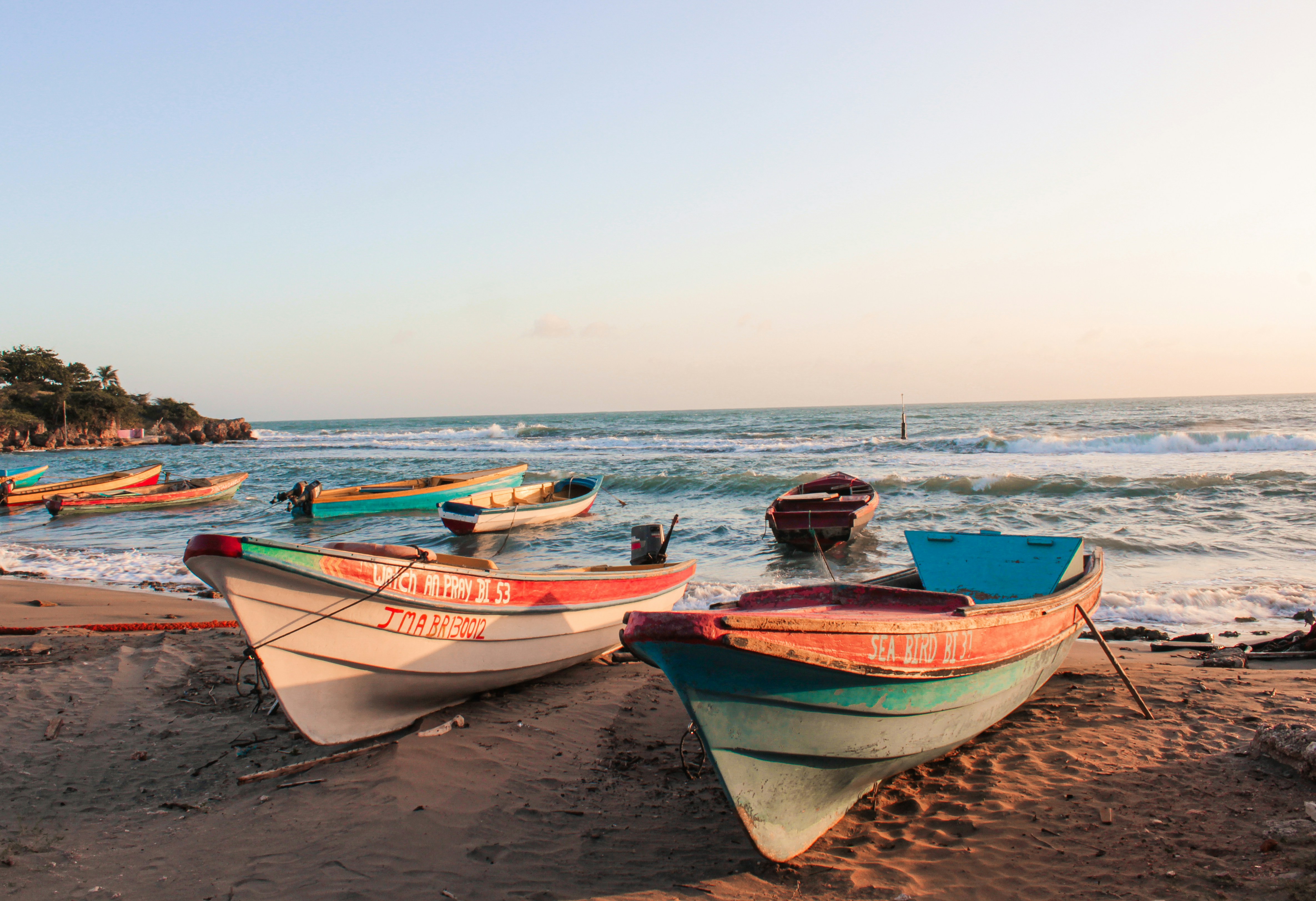 Several colorful wooden fishing boats, painted in shades of red, white, and turquoise, are resting on a sandy beach at sunset. Gentle waves roll in from the Caribbean Sea under a pale, clear sky.