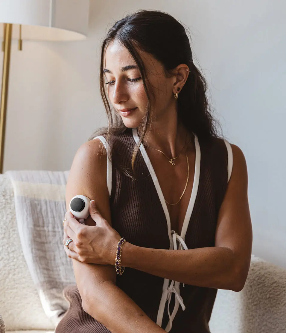 Woman using a Rythm at-home blood collection device on her upper arm, seated indoors in soft natural light, looking down as she applies the device.