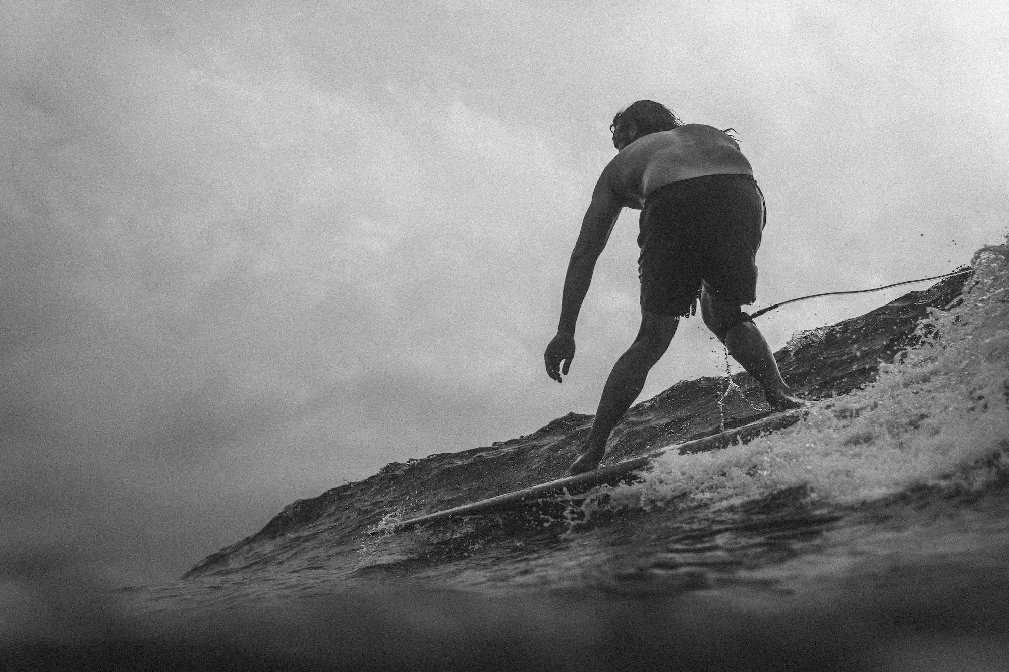 A silhouette of a surfer navigating a wave against a cloudy sky in a black and white photograph.