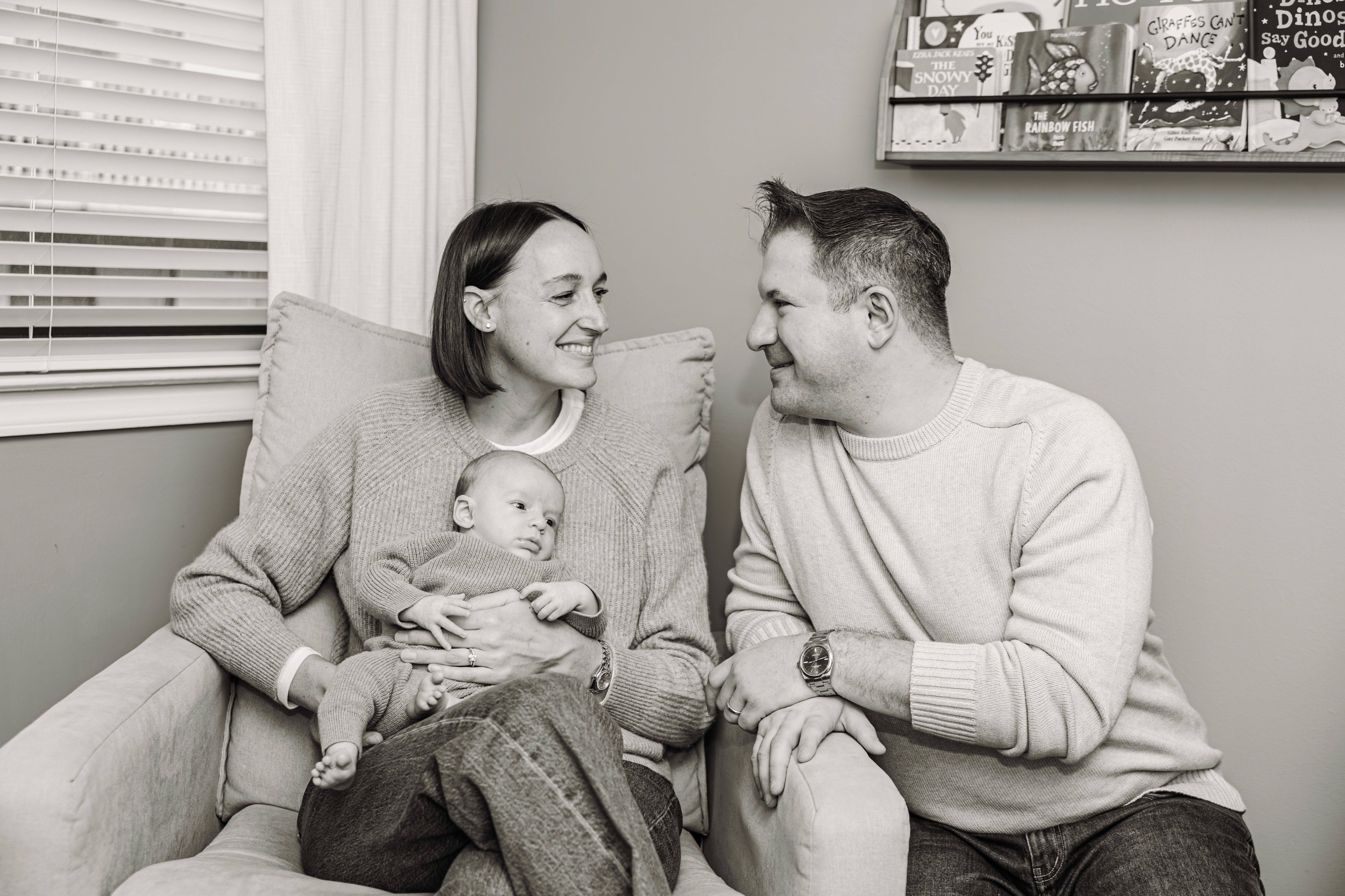 New parents sharing a tender look with their newborn nestled between them at home in White Plains, NY — intimate, emotional black and white newborn and family photography by Lizz Spano Photography, Westchester County newborn photographer.
