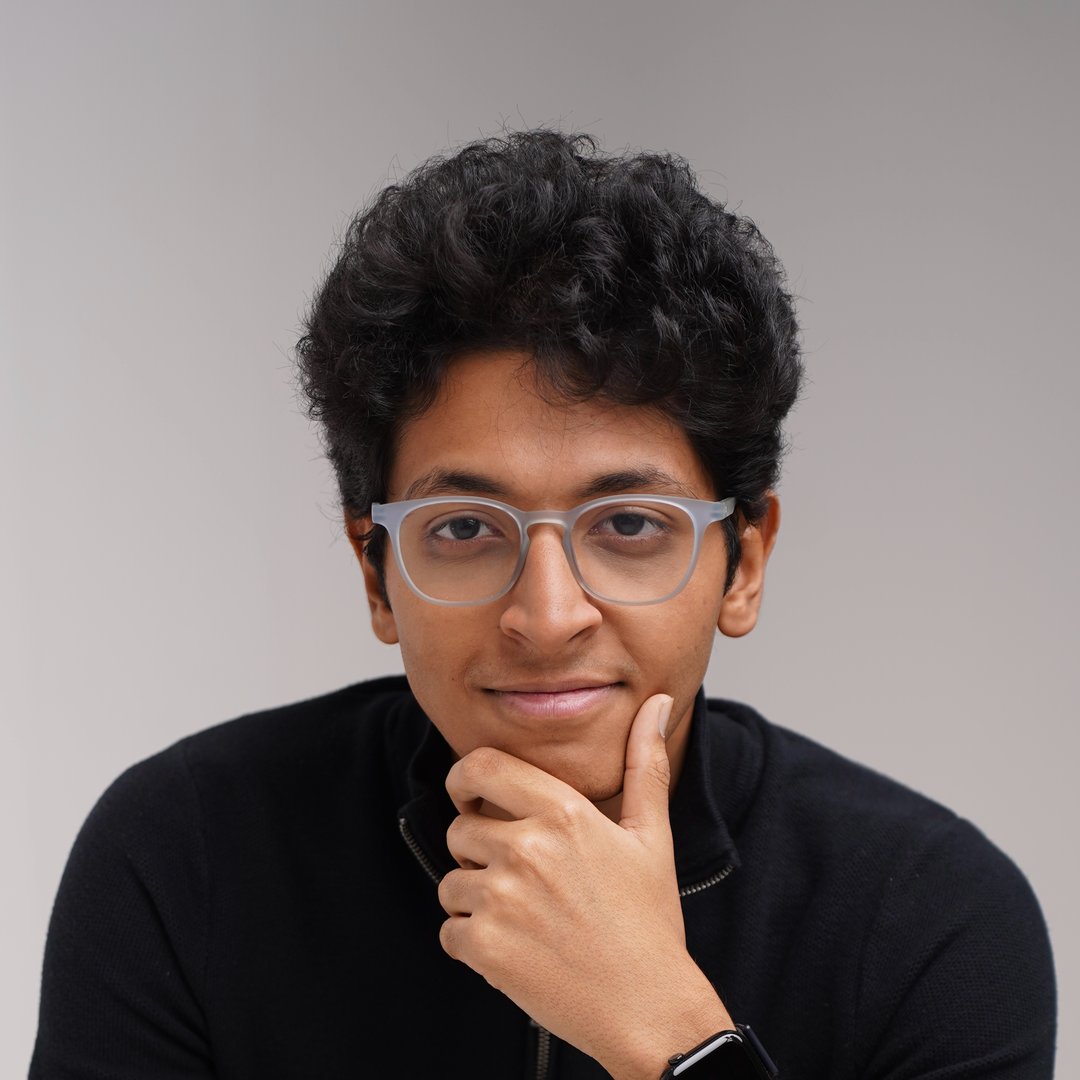 A woman with blonde bob hair in a grey coat and beige turtleneck stands against a neutral backdrop. She has a serious expression, conveying elegance.