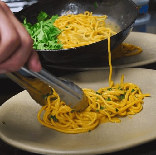 Chef using tongs to plate freshly cooked spaghetti with herbs from a pan onto beige dishes.