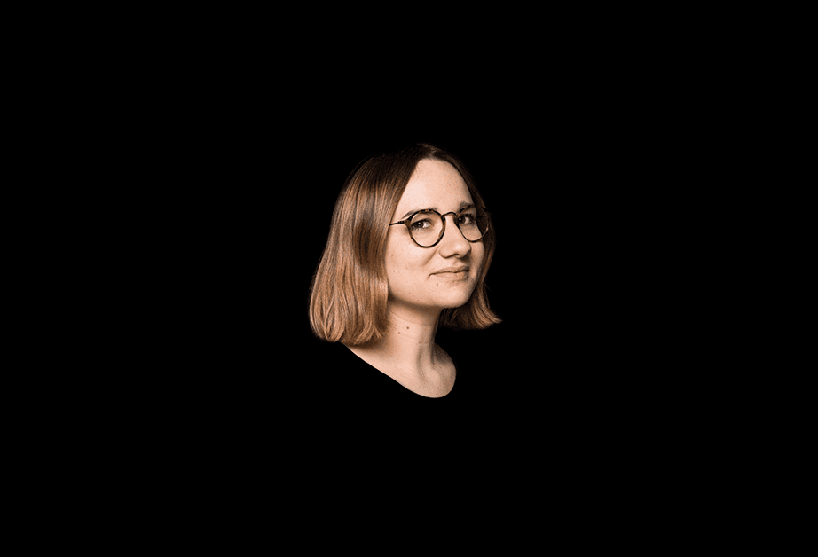 A woman with shoulder-length light brown hair and round, dark-framed glasses is shown in profile against a solid black background. She is wearing a dark-colored top and looking toward the right side of the frame.