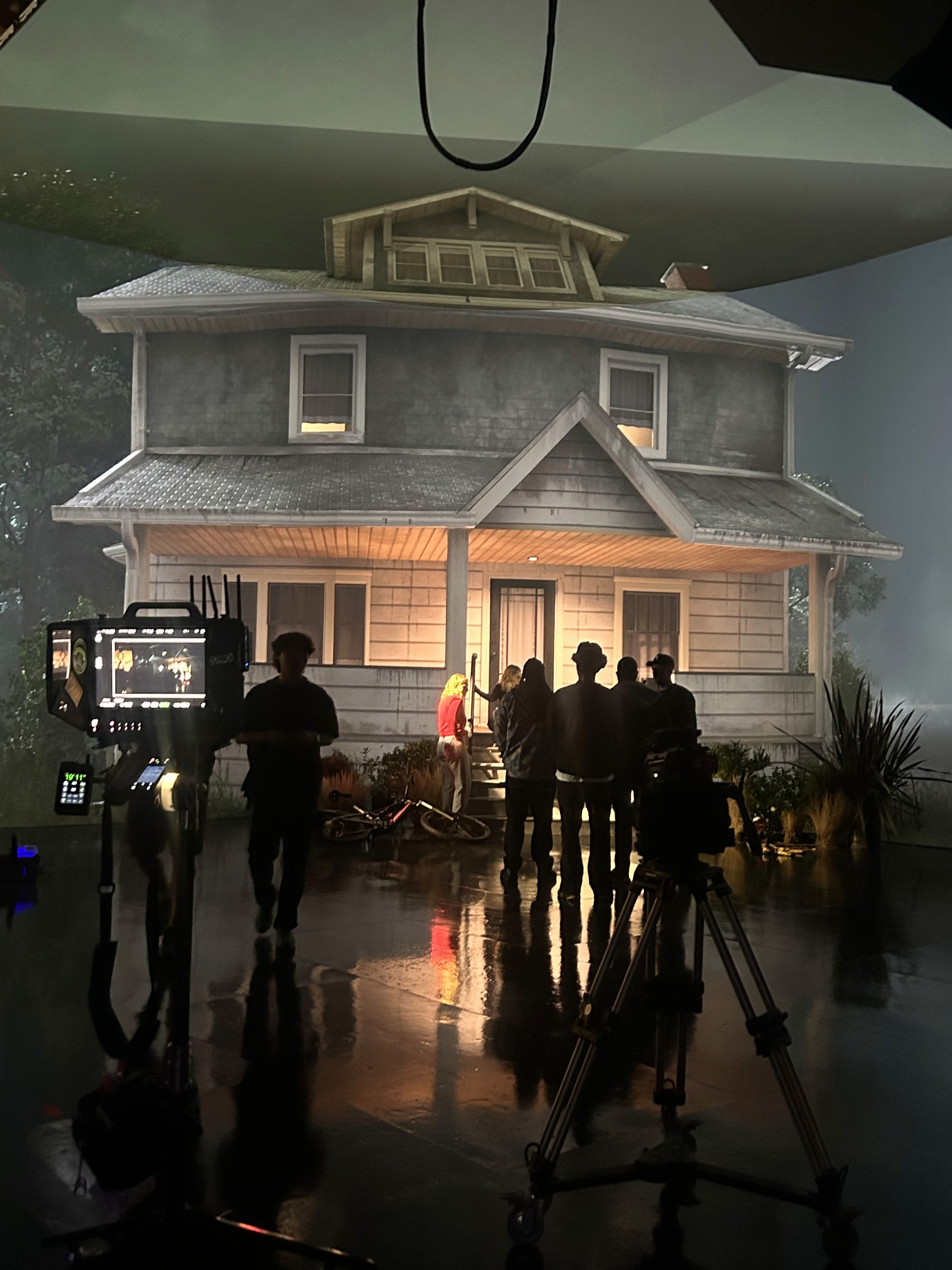 A live-action production crew films on a virtual production stage featuring a two-story house rendered in Unreal Engine. The house, displayed on a large LED volume wall, is lit to mimic dusk or early evening. Crew members gather around a camera setup in the foreground, with lighting equipment and monitors visible, capturing the seamless integration of real actors and digital environments.