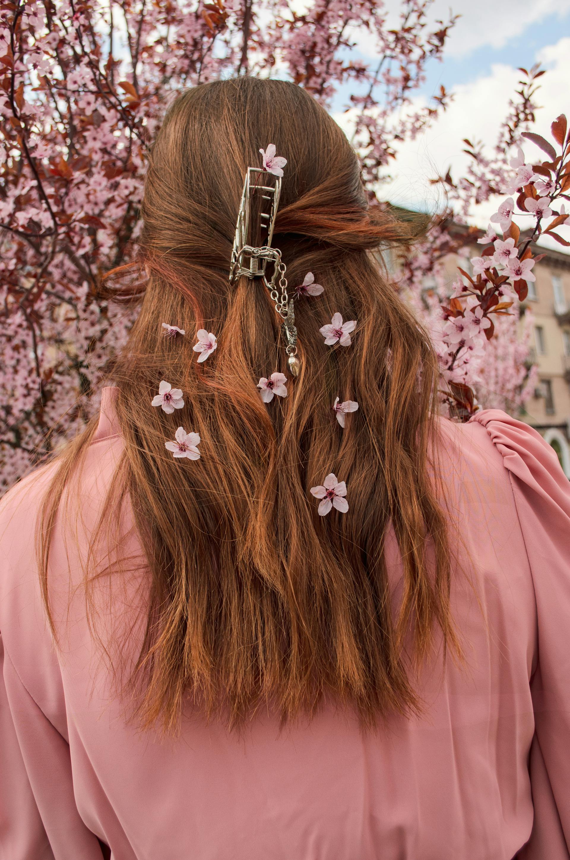 The back of a woman with long auburn hair held by a silver claw clip, decorated with several small pink cherry blossoms and standing in front of a blooming tree.