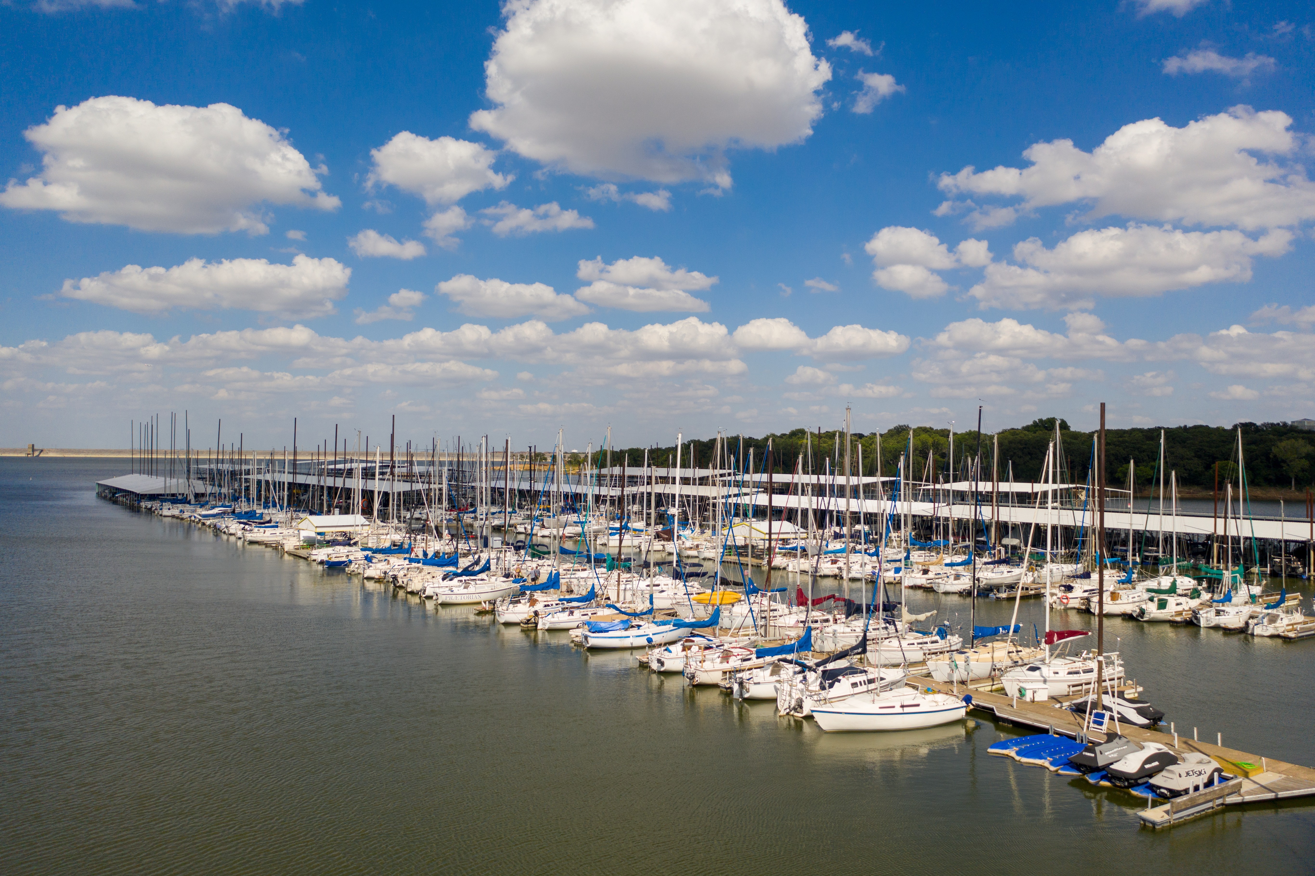 A serene marina filled with numerous sailboats and yachts docked in calm water under a bright blue sky dotted with fluffy white clouds.