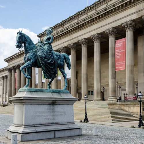 대형 기둥과 현수막이 있는 웅장한 건물 앞에 있는 청동 승마 조각상. 전경에는 자갈이 깔린 산책로가 보입니다.
