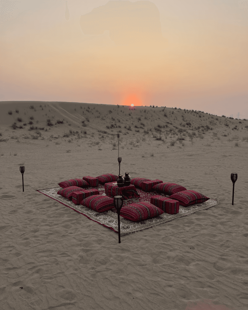 Two softly lit canvas tents on the desert at dusk, viewed from inside another tent with camping chairs visible outside.