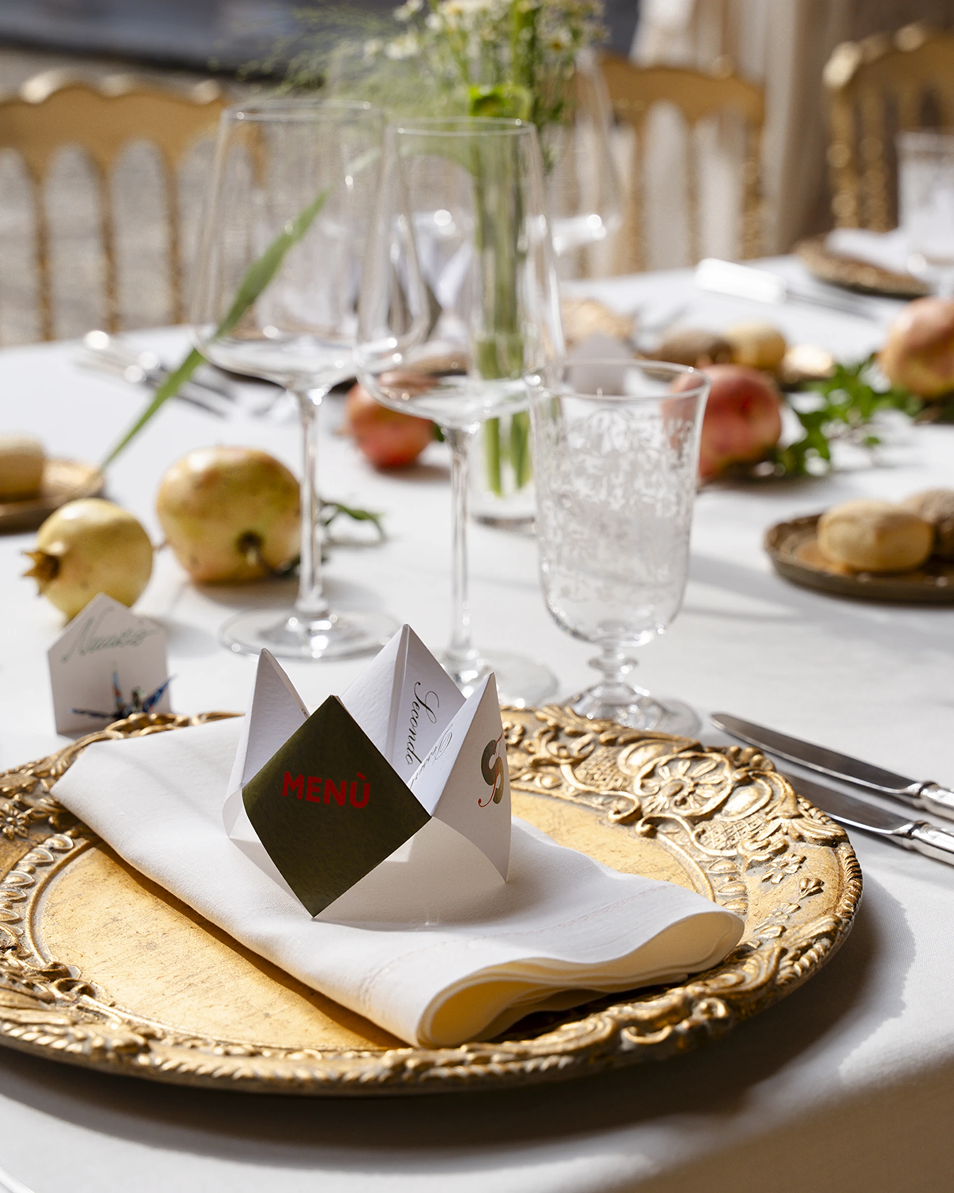 Dettaglio di un piatto dorato con tovagliolo di lino e menu piegato a origami, tra bicchieri di cristallo e melograni