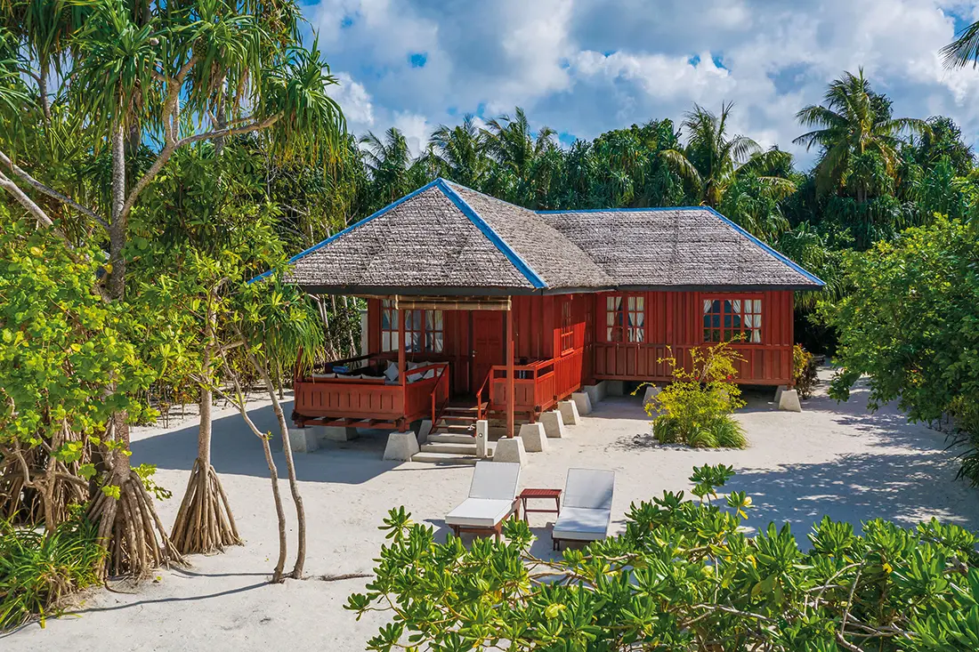 Ein klassischer Strandbungalow aus rötlichem Holz mit Reetdach, der auf kleinen Betonpfeilern im weißen Sand steht, umgeben von tropischen Bäumen und zwei Sonnenliegen im Vordergrund.