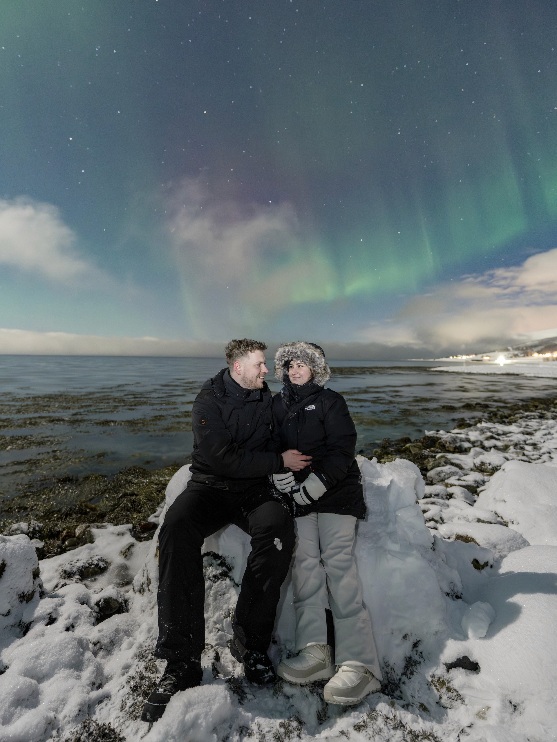 Couple seated closely on the shore, looking at one another beneath a subdued aurora and starry sky.