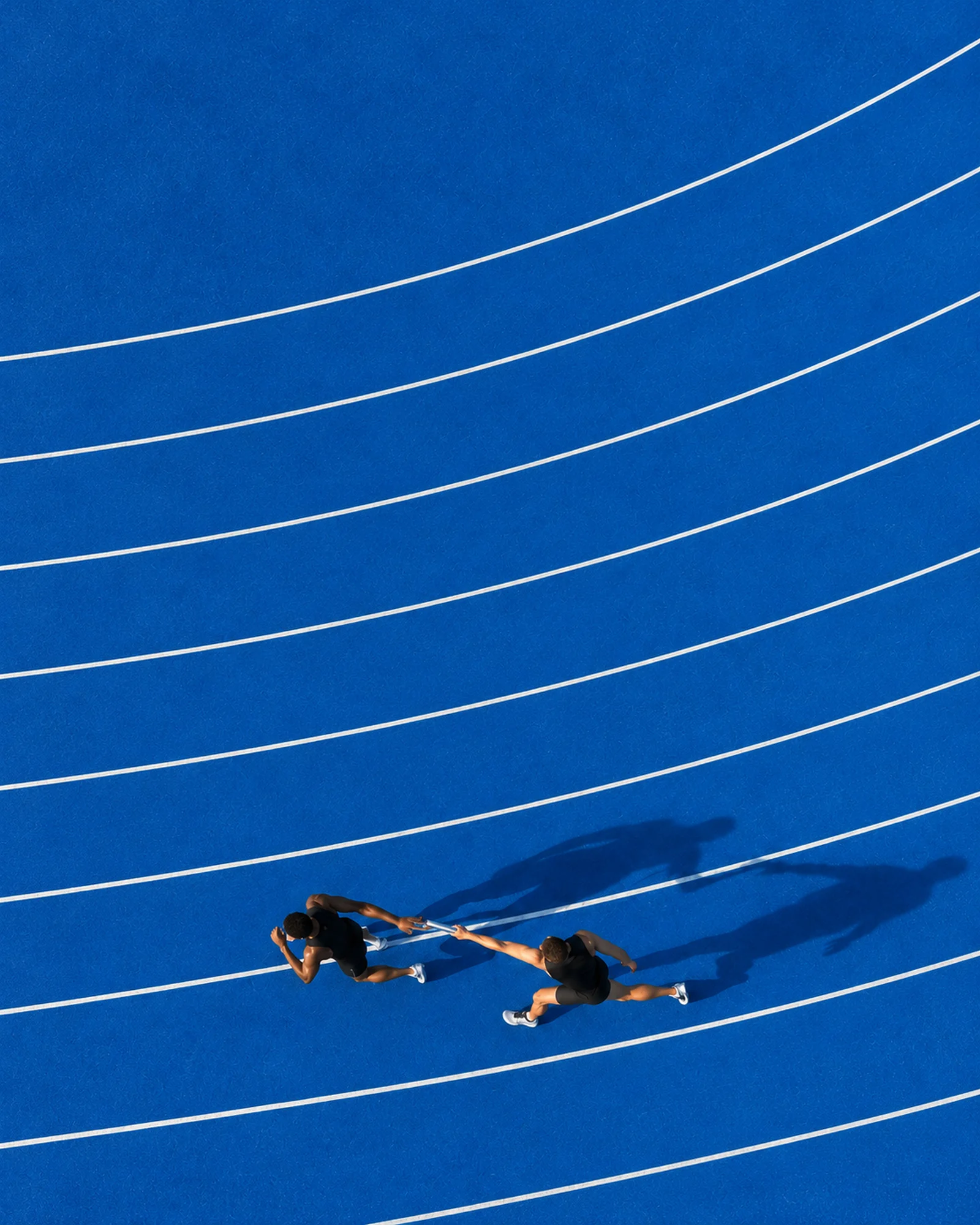Two relay runners passing a baton on a blue running track, seen from above