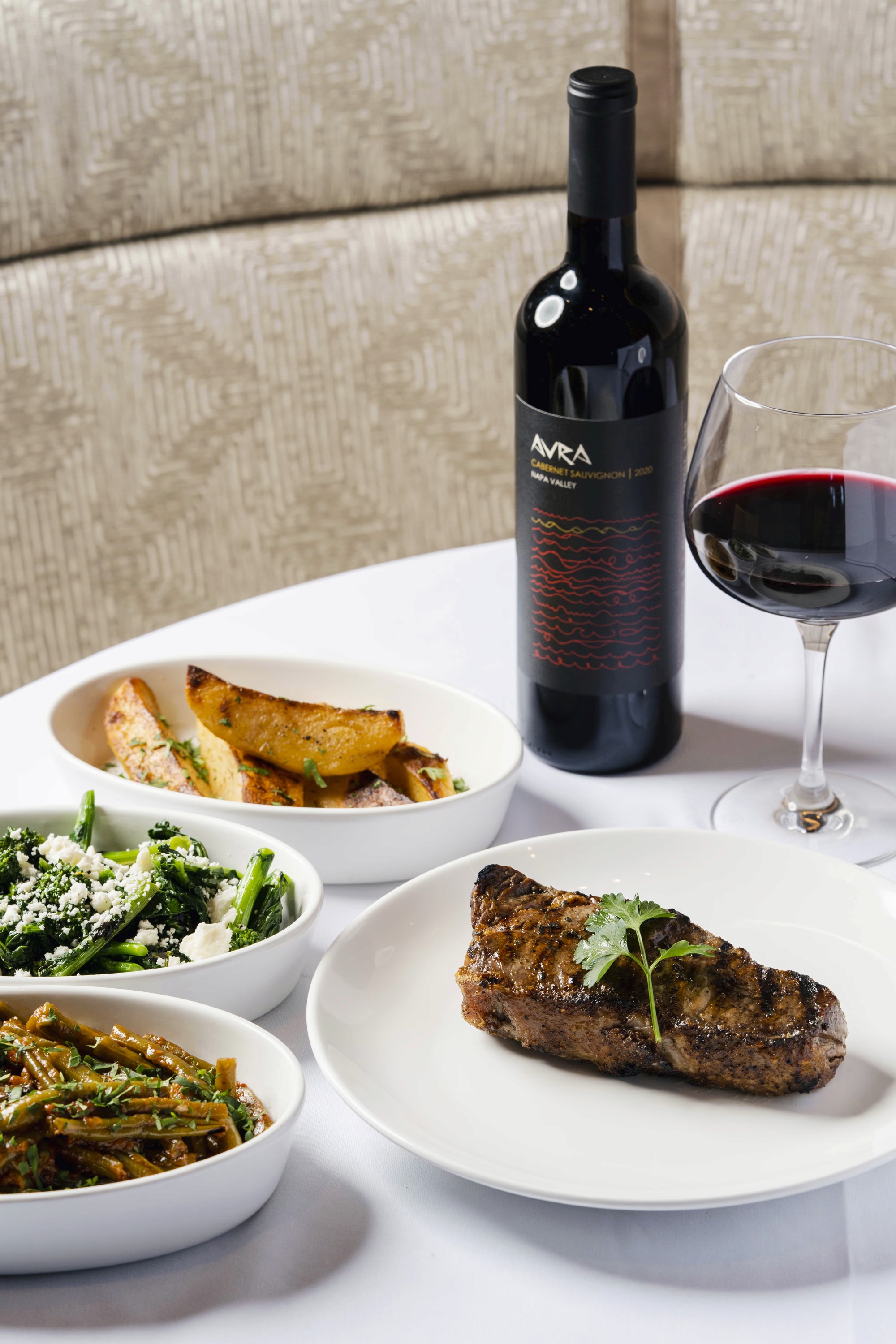 A table set with a bottle of red wine, a glass, a plate of steak, and bowls of vegetables and bread.
