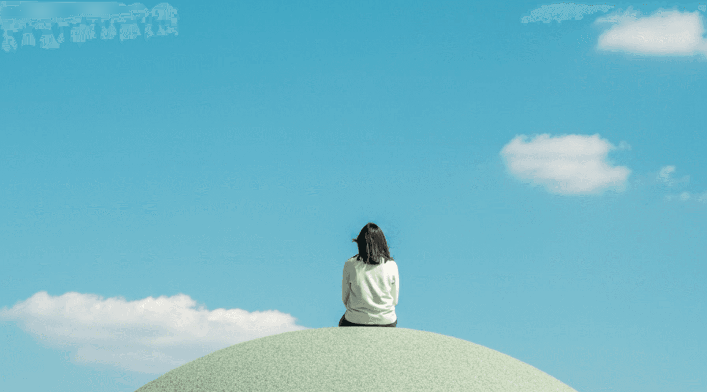 A solitary figure sitting on a rounded grassy dome, gazing at a clear blue sky dotted with a few white clouds, creating a serene and contemplative atmosphere.