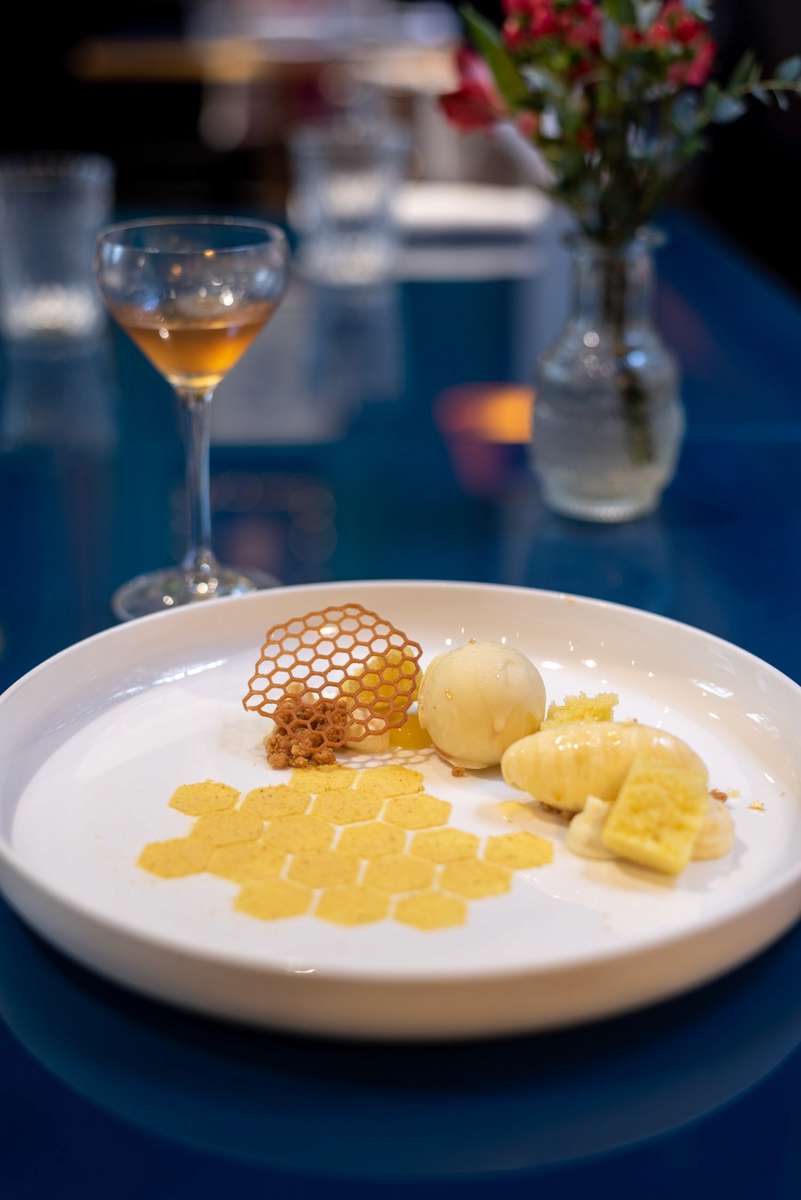 A plated dessert with yellow and white components and a chocolate cage sits before a glass of amber liquid on a blue table.