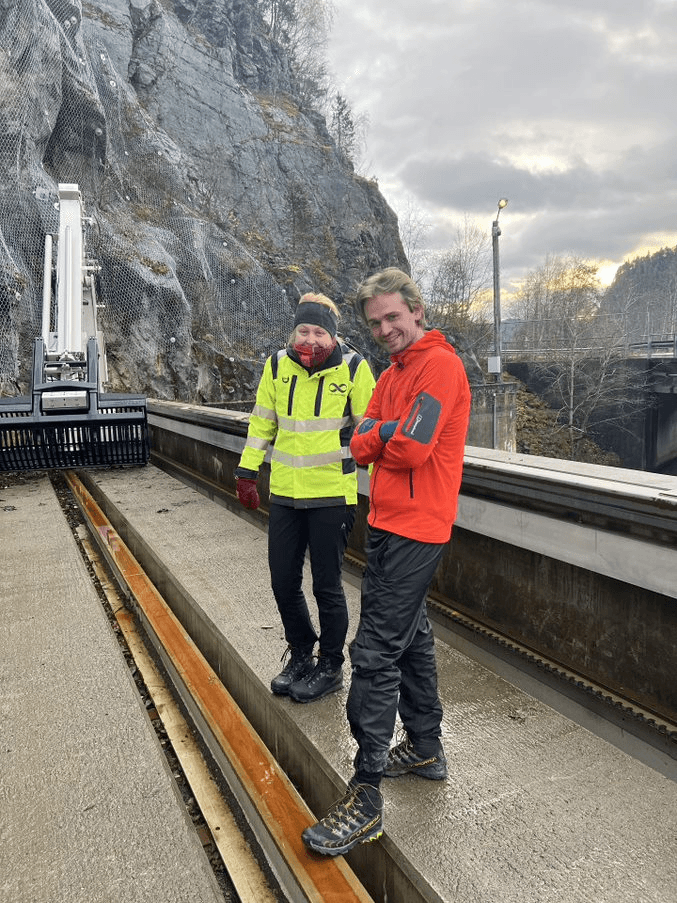 A person in an Aneo yellow jacket and a male trainee are conducting an inspection in front of the entrance to a hydropower plant.