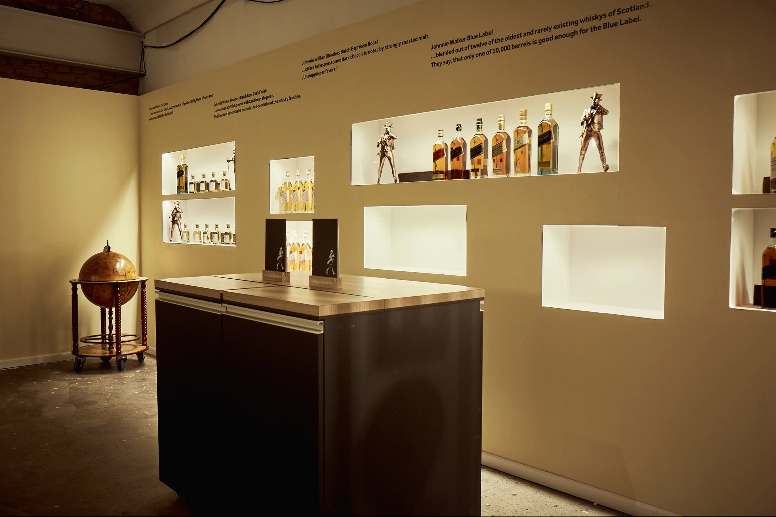 Johnnie Walker exhibition wall at DIAGEO BCB17 with illuminated niches holding whisky bottles and striding-man figures, a vintage globe bar and a dark counter.