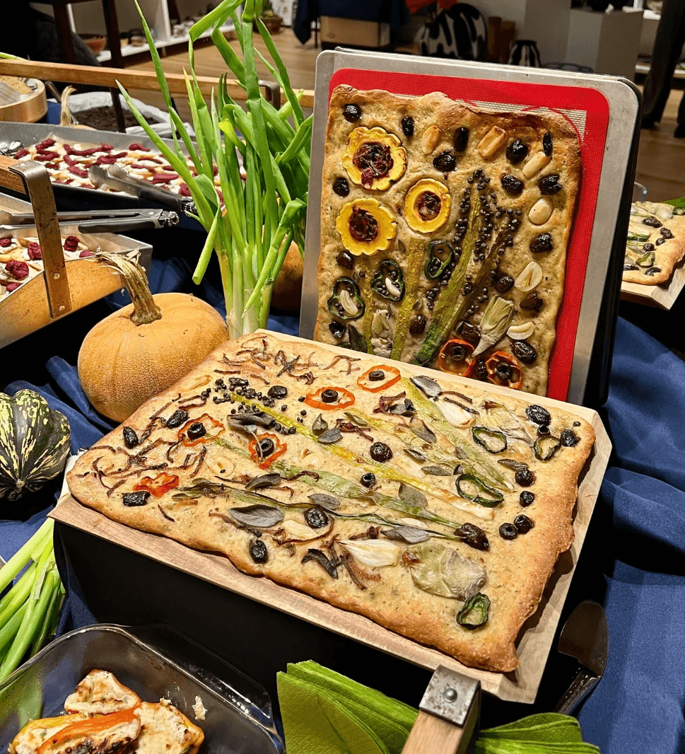 Seasonal vegetable focaccia topped with squash, peppers, onions, and herbs displayed at a community gathering.
