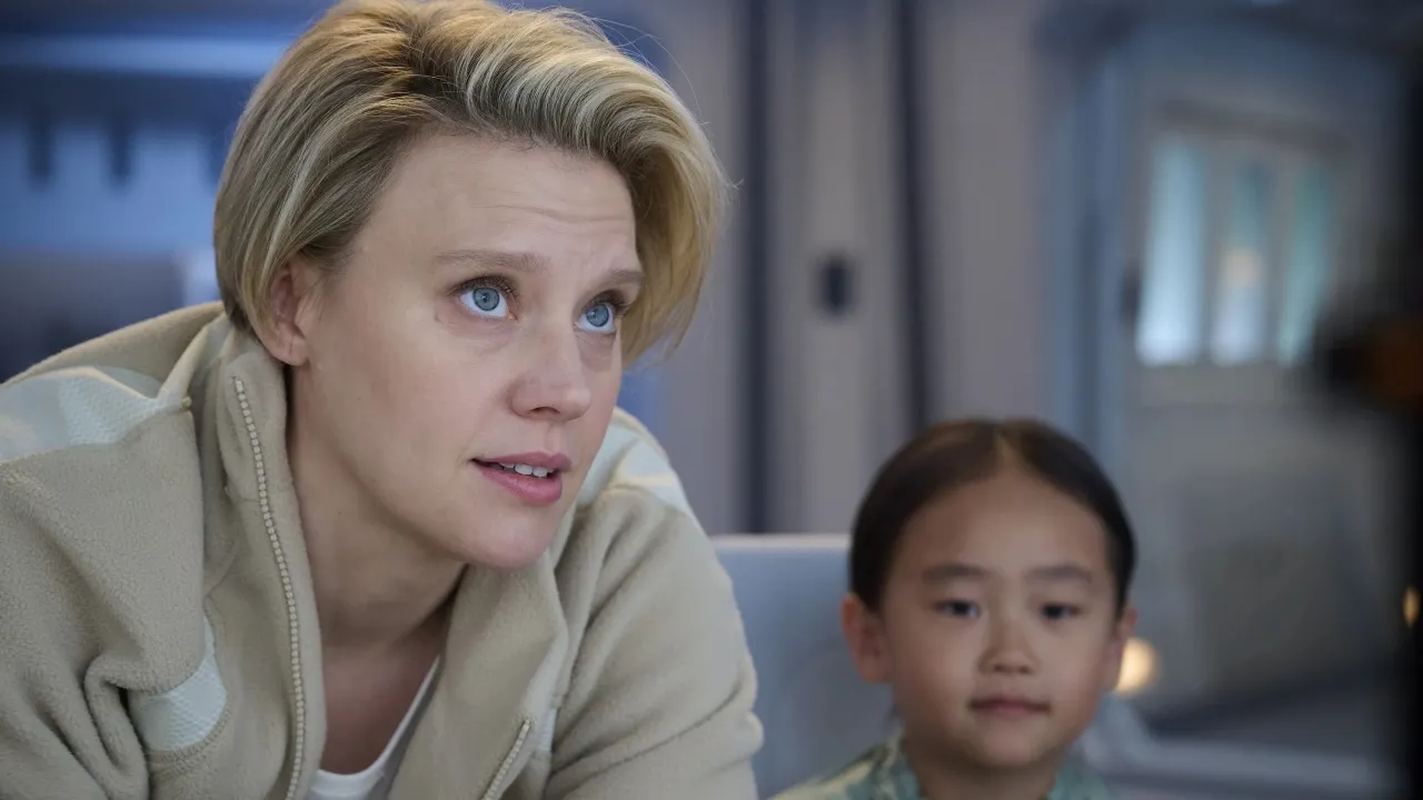 A woman with short blond hair looks attentively ahead, sitting next to a child in a softly lit futuristic room, creating a focused and thoughtful atmosphere.